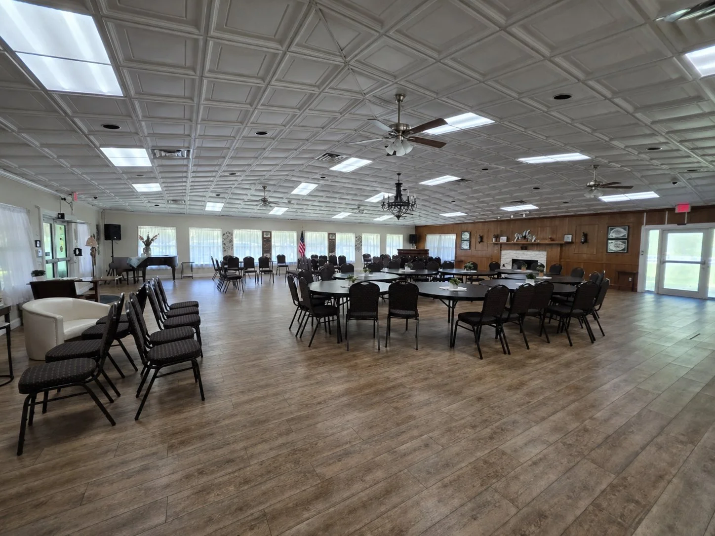 Large banquet hall with multiple round and rectangular tables, black chairs, wood paneling on one wall, a fireplace, and windows along the outer wall. The ceiling has decorative tiles, ceiling fans, and fluorescent lighting.