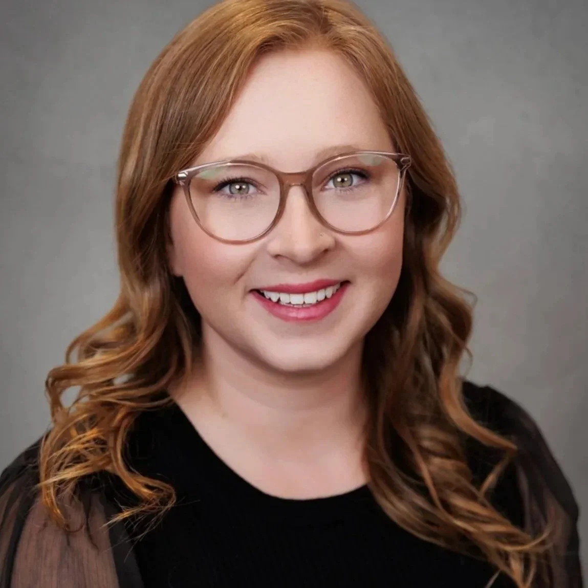 A young woman with shoulder-length black hair smiling indoors surrounded by green plants.