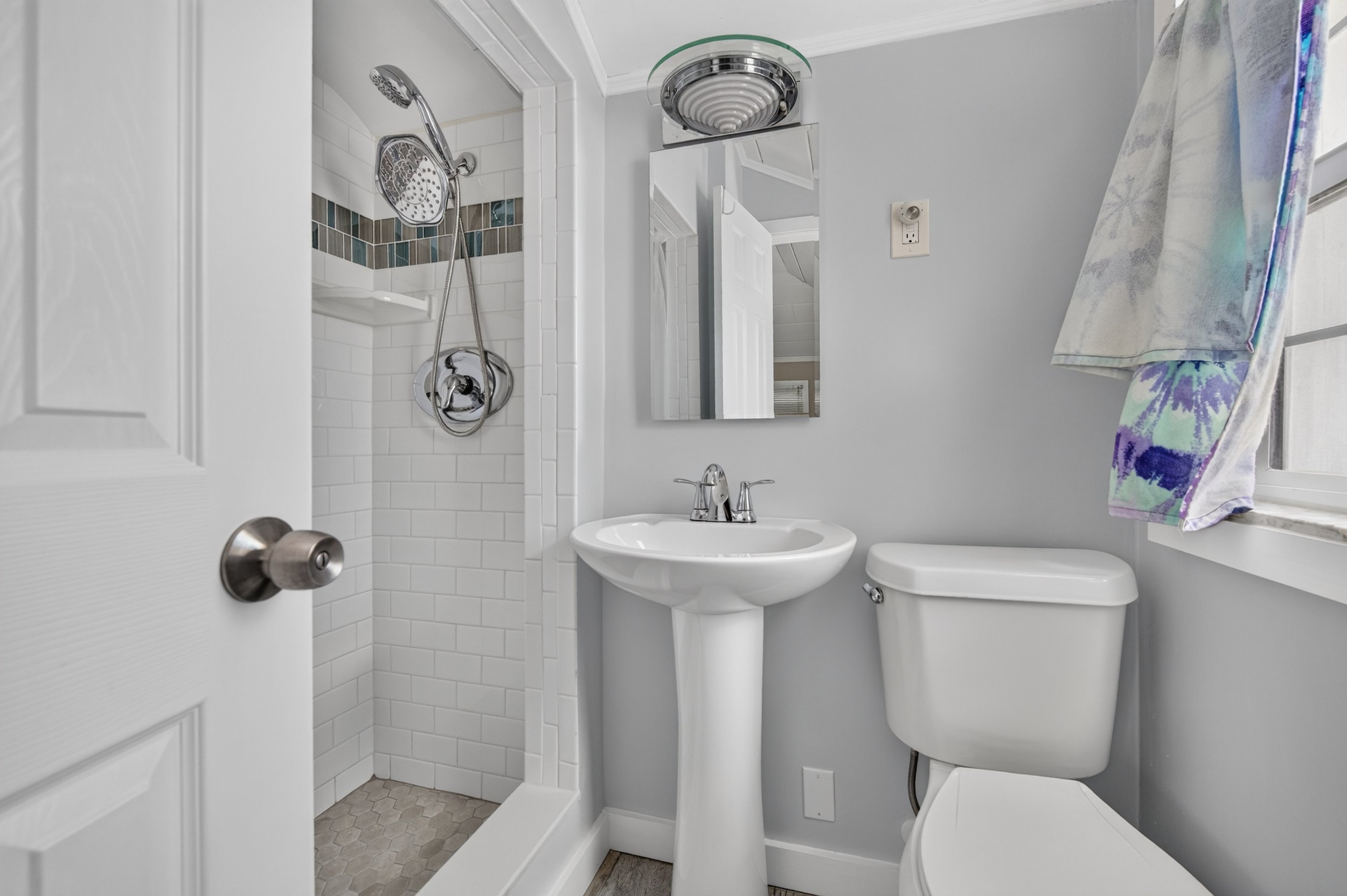Small bathroom with white tiles in the shower area, showerhead and controls, small sink with chrome faucet, toilet, mirror above the sink, window with a floral towel hanging, light gray walls, and beige hexagonal floor tiles.