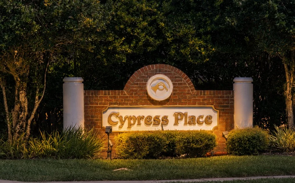 Residential entrance sign reading 'Cypress Place' with brick background, topped with a circular emblem, flanked by white columns, surrounded by bushes and trees at dusk.