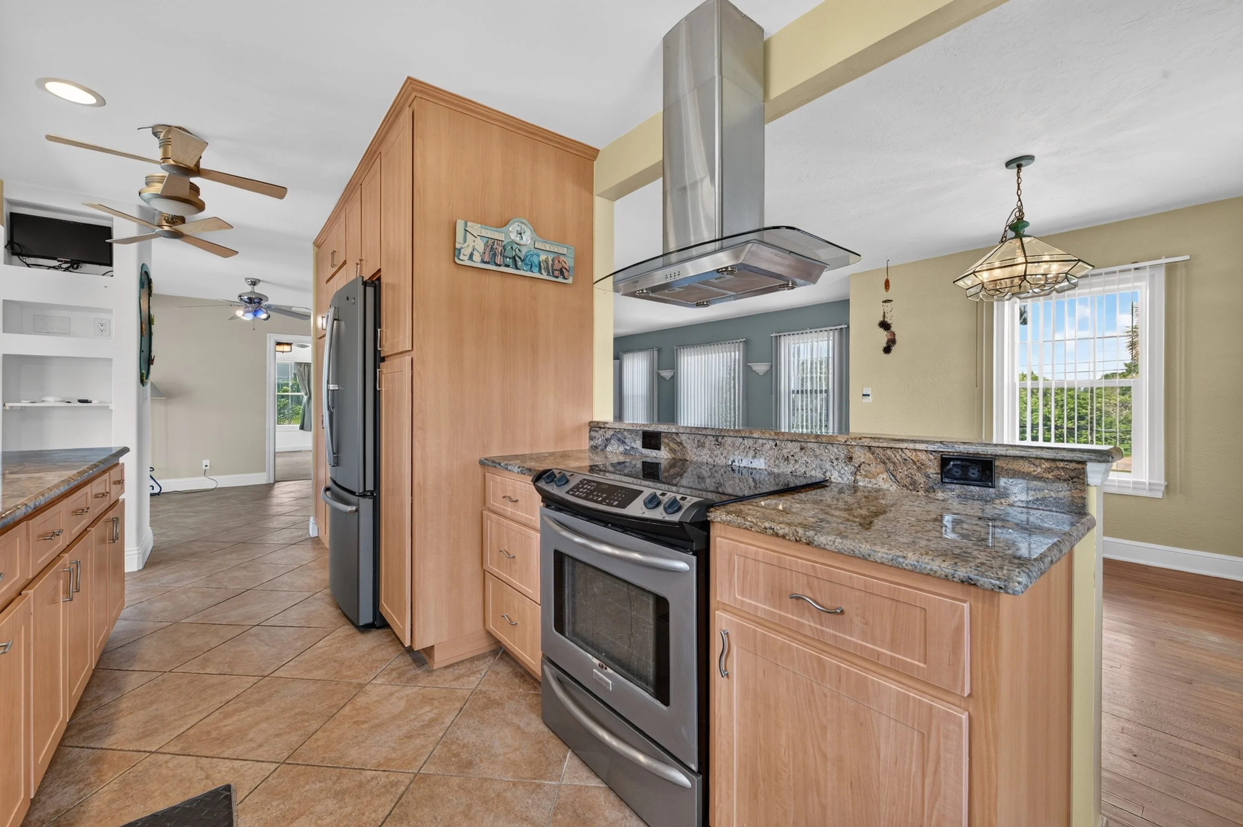 A kitchen with wooden cabinets, a stainless steel stove, granite countertops, and large windows with vertical blinds, and a ceiling fan.