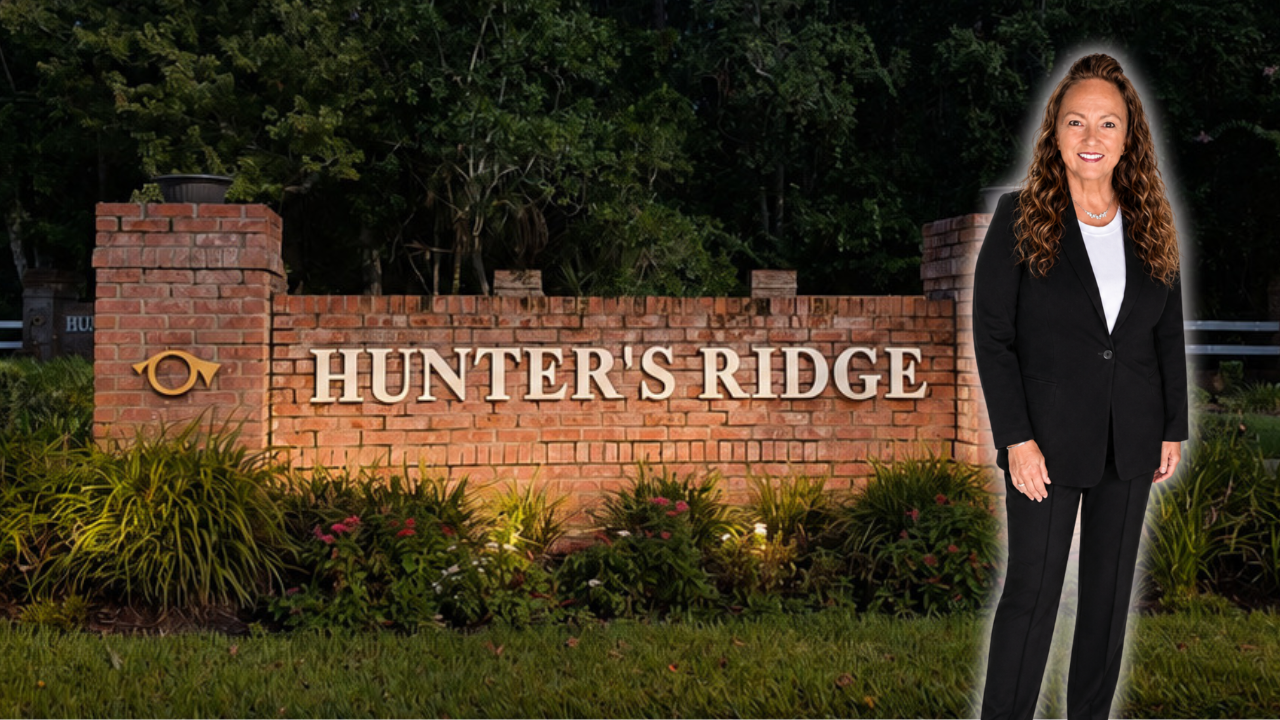 A woman with long curly hair in a black suit standing in front of a brick sign that reads 'Hunter's Ridge' at dusk, with greenery and trees in the background.