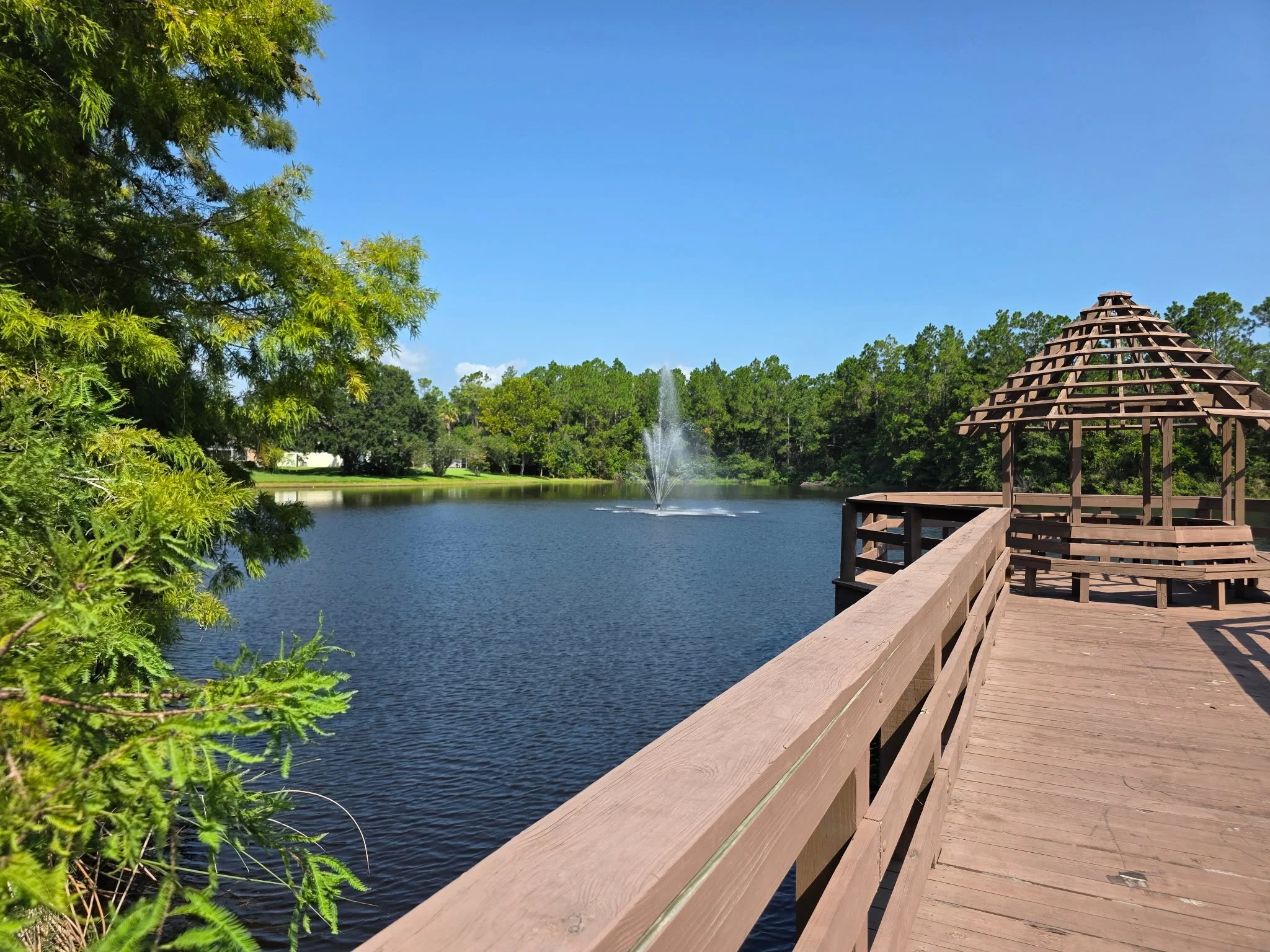 A wooden observation deck extending over a lake with a fountain, surrounded by lush green trees under a bright blue sky.