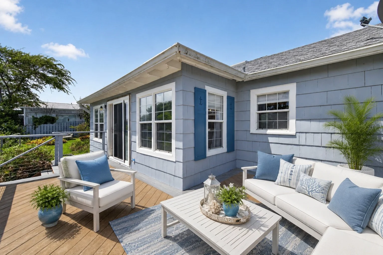Outdoor patio with white seating and blue cushions, potted plants, and a view of a house with blue siding and white trim, sunny weather, and a partly cloudy sky.