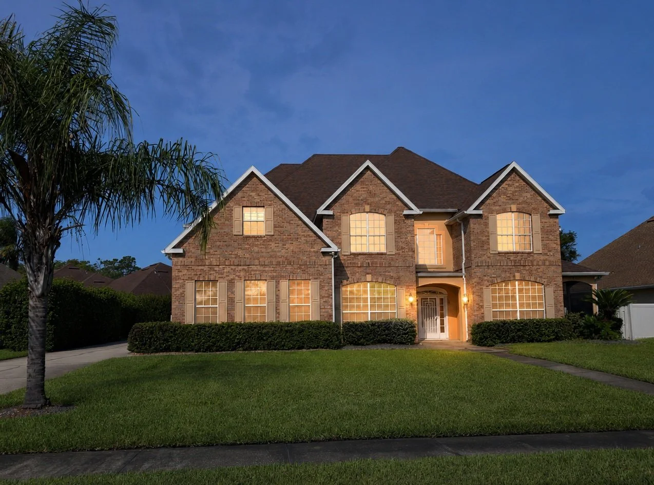 A two-story brick house illuminated by warm exterior lights during dusk, with a front lawn, walkway, and a palm tree on the left side.