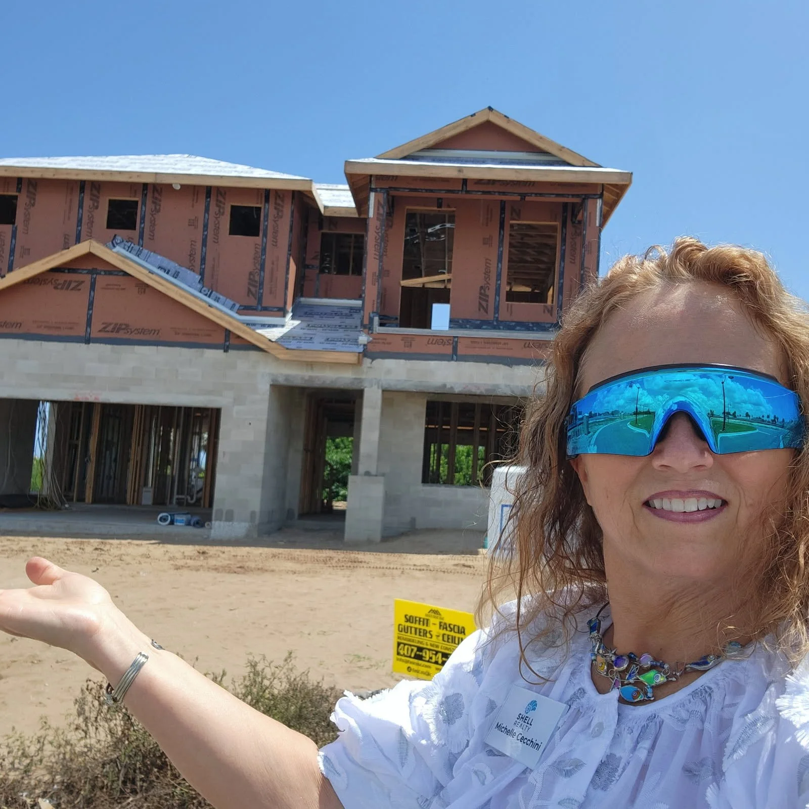 A woman in safety glasses and a white shirt with a name tag standing in front of a house under construction, pointing towards it.