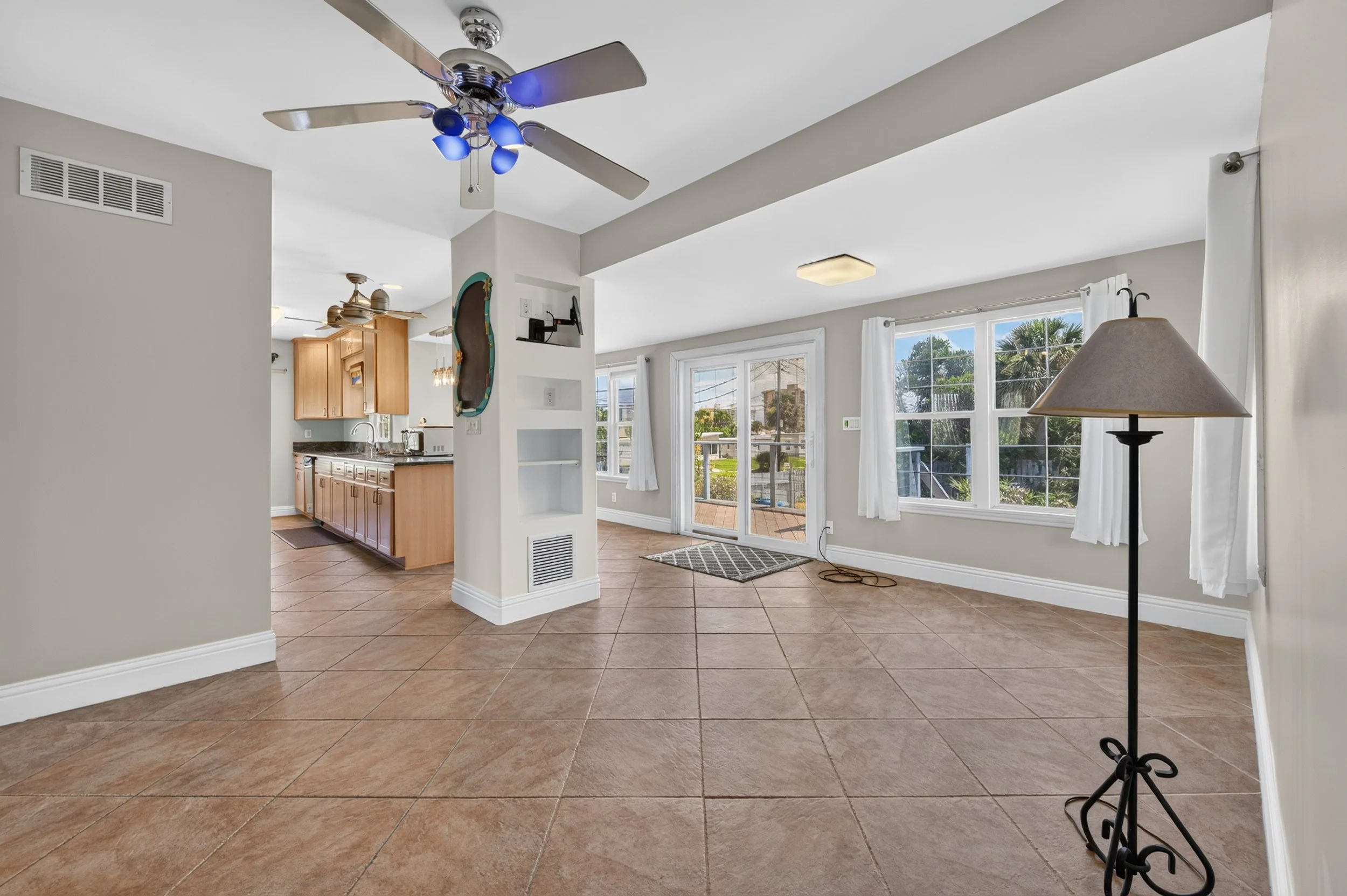 Interior of a bright living room with large windows, tan tiled floor, white walls, ceiling fans, a standing lamp, and a view of an outdoor patio and garden.