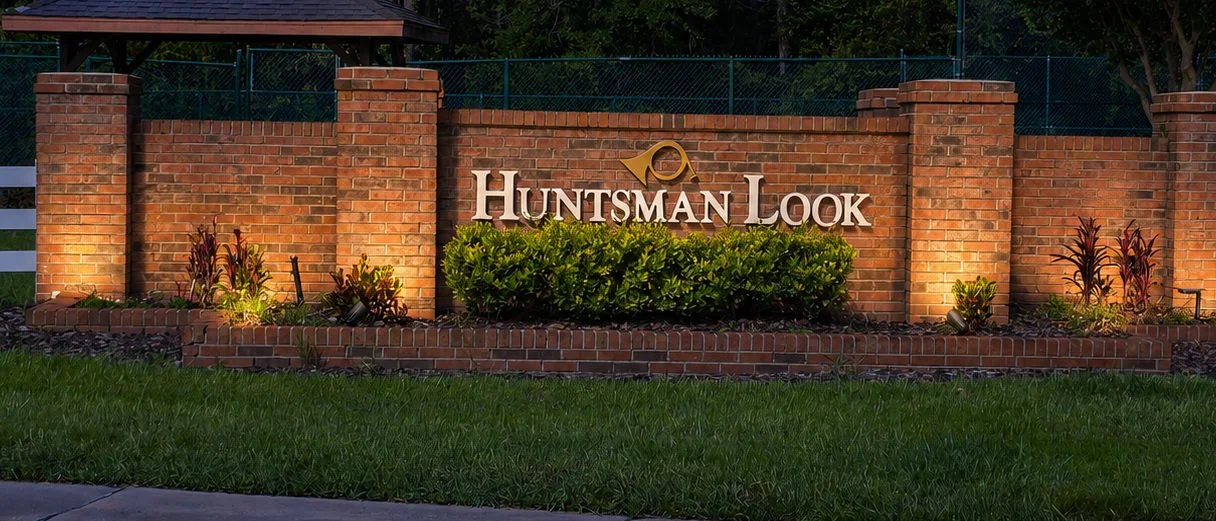 A brick sign with the words "Huntsman Look" and a magnifying glass logo, surrounded by landscaped plants and lit with lighting, at dusk or evening.