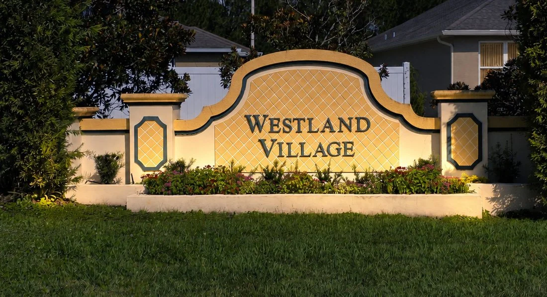 A sign that reads 'Westland Village' with decorative plants and flowers in front, surrounded by trees and residential houses.