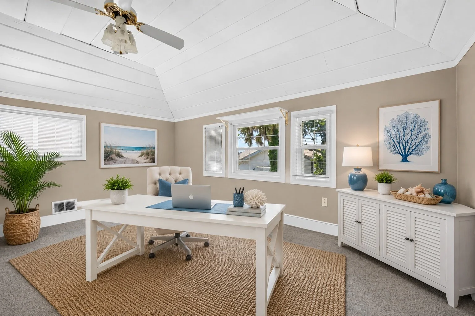 Bright home office with white desk, beige walls, and multiple windows with blinds, decorated with coastal-themed artwork and decor, including a large potted plant, blue vases, and seashells.