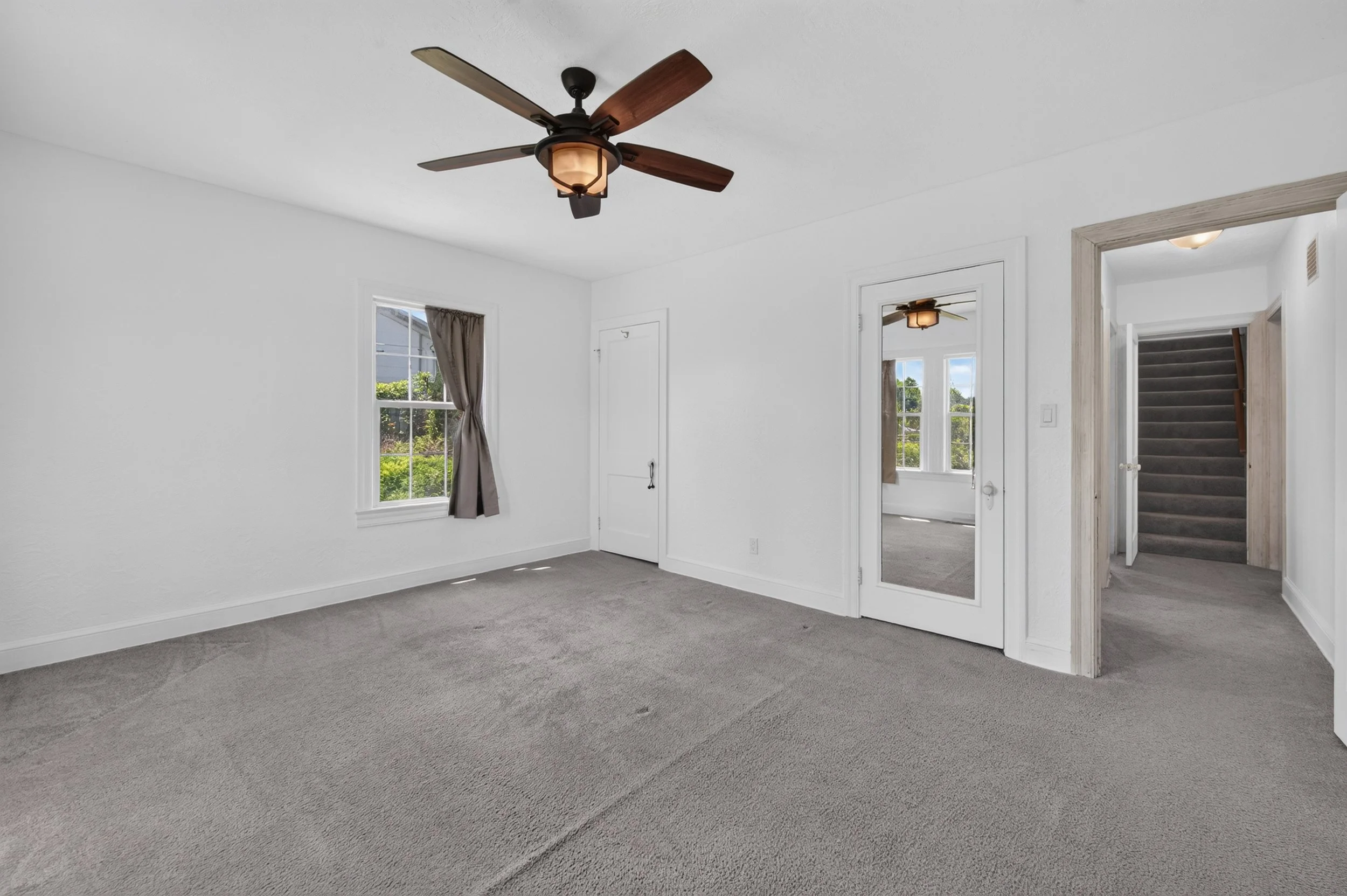 Empty living room with white walls, gray carpet, ceiling fan, two windows with curtains, and a doorway leading to a staircase.