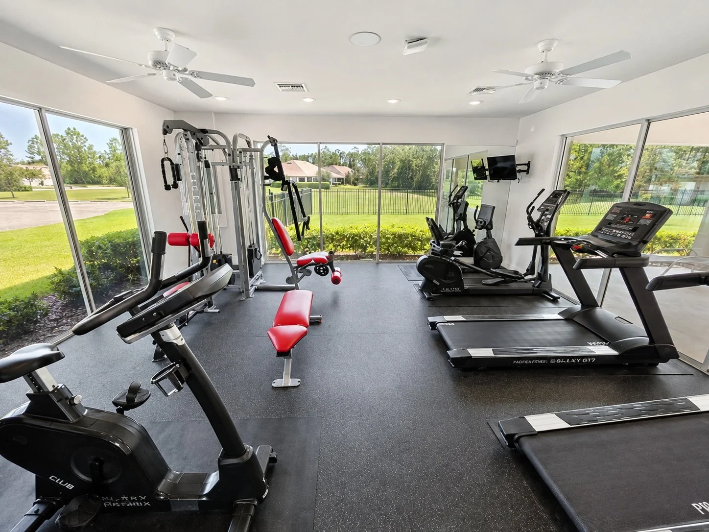 Gym room with treadmill, exercise bike, weight machine, and red and black weight bench, large windows showing greenery outside.