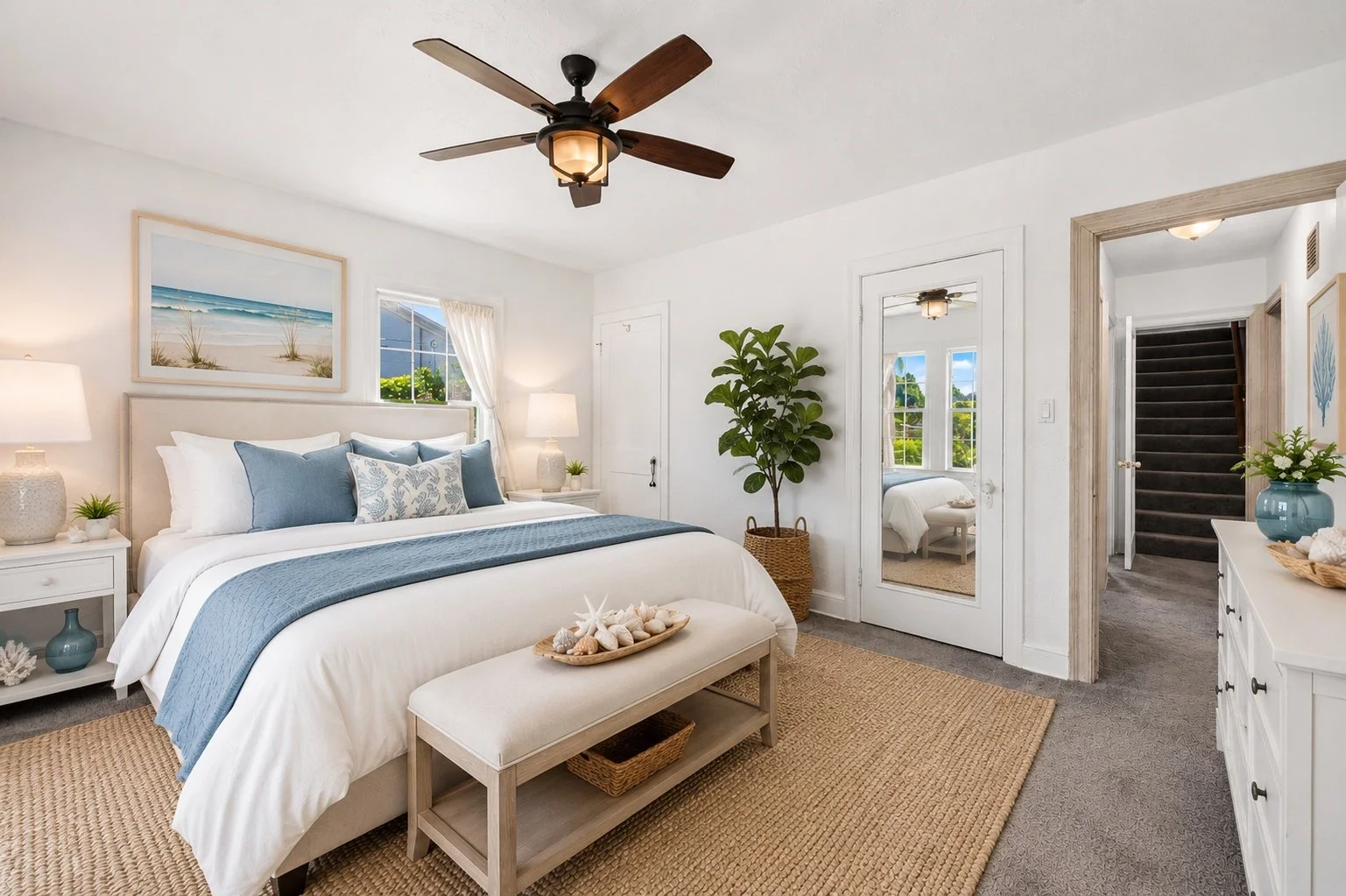 A bright coastal-themed bedroom with a white bed, blue and white pillows, a wooden bench, a large houseplant, and decor inspired by the beach, including seashells and starfish, with natural light coming through the windows.