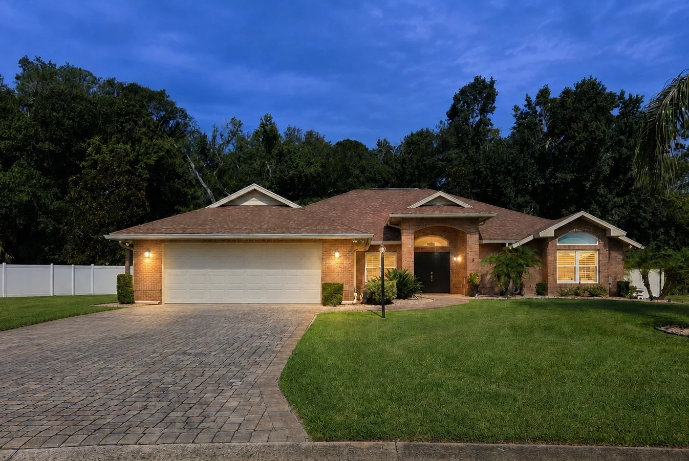A cozy brick house with a two-car garage, front yard, and lush greenery at dusk with outdoor lights on.