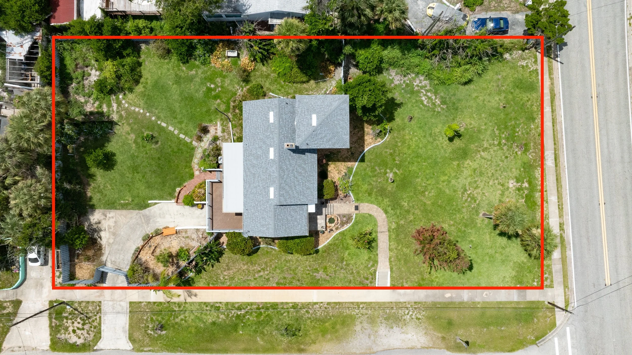 Aerial view of a house with a gray roof, surrounded by a large, green yard with trees and a paved walkway, bordered by a street with parked cars.