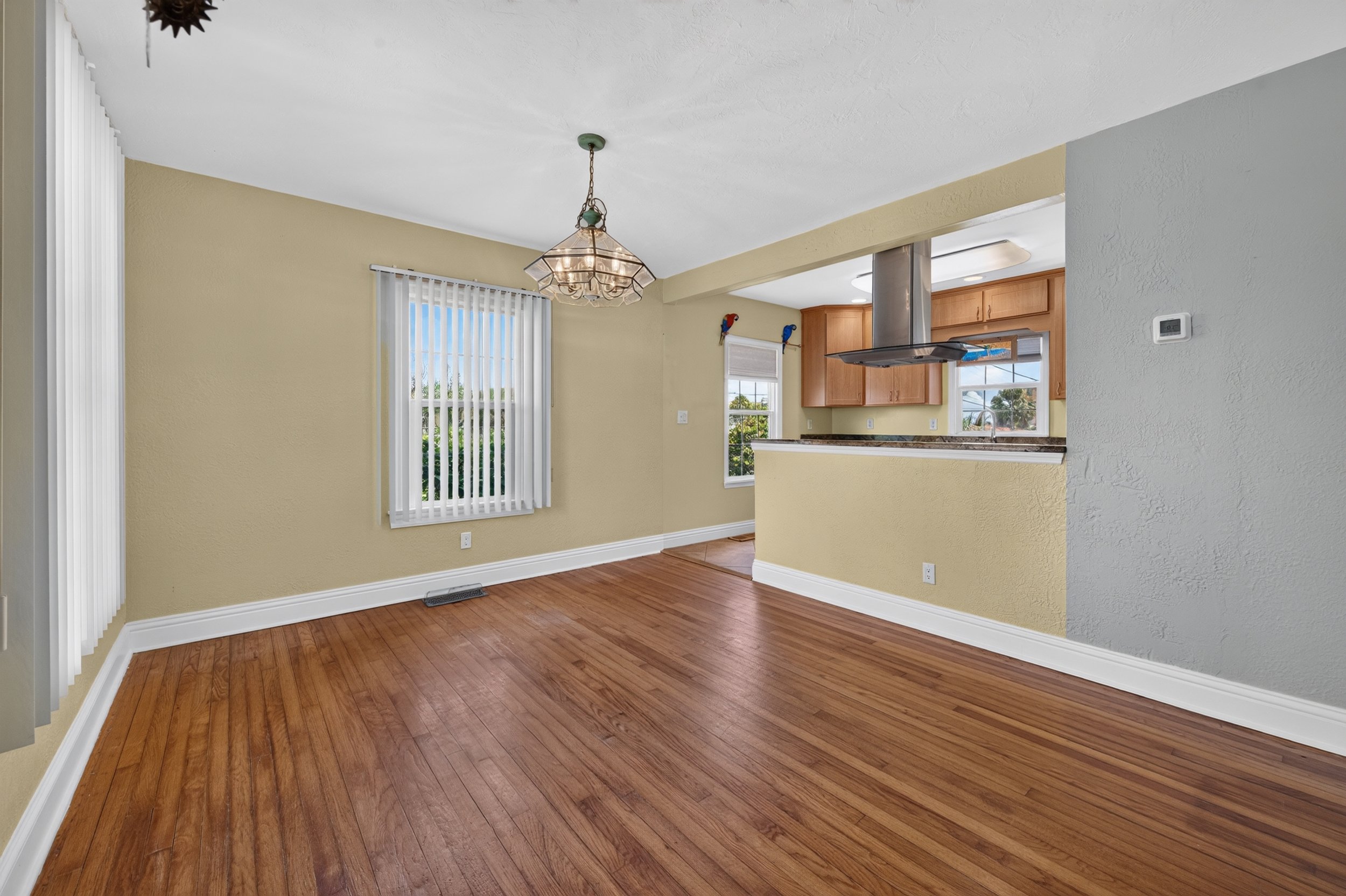 An open concept kitchen and dining area with hardwood floors, yellow walls, white trim, a chandelier, a window with vertical blinds, and a partial view of the kitchen cabinets and range hood.