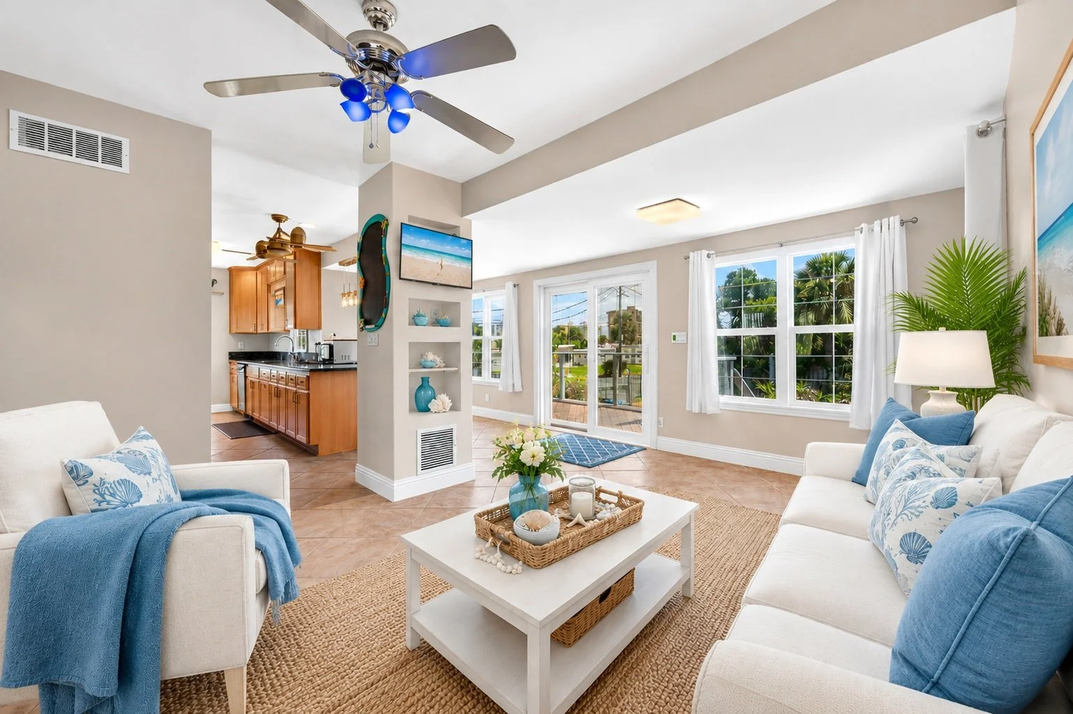 Bright living room with white couches, blue pillows, and a coffee table with nautical decorations, adjacent to a kitchen with wooden cabinets, and large windows showing outdoor greenery.