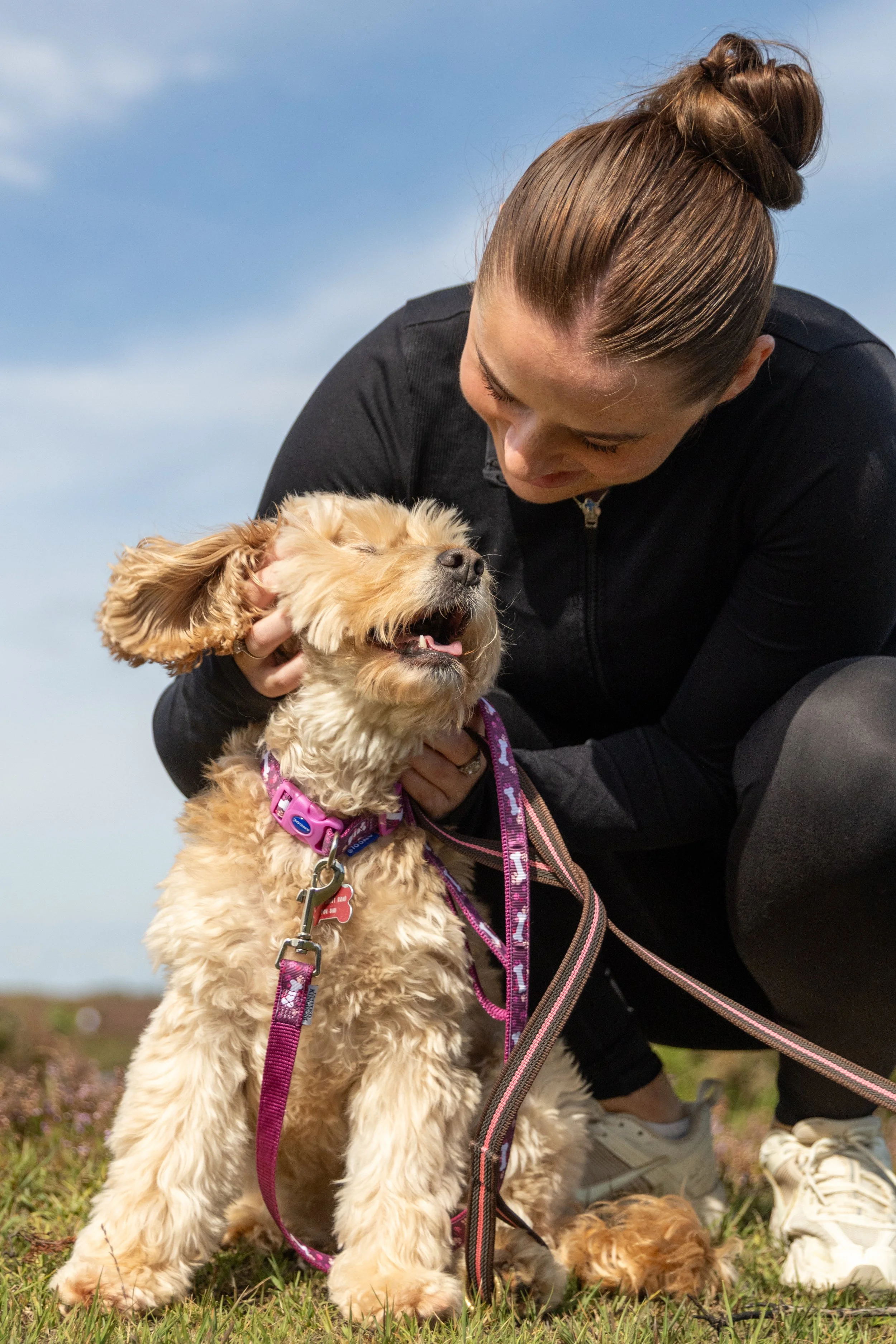 A young dog trainer woman with brown hair in a bun smiling as she pets a fluffy, tan-colored puppy, cockapoo,  with floppy ears outdoors on grass during the daytime. Reactivity Dog Training