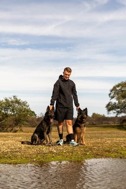 A dog trainer in black athletic clothing with two fully trained German Shepherd dogs outdoors near a body of water, with trees and a cloudy sky in the background, in the New Forest, Hampshire