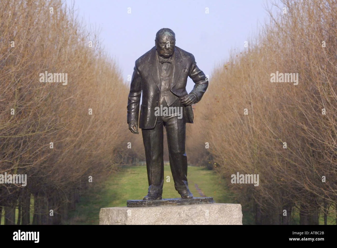 Bronze statue of Winston Churchill in a suit standing on a stone pedestal in an outdoor park with leafless trees in the background.