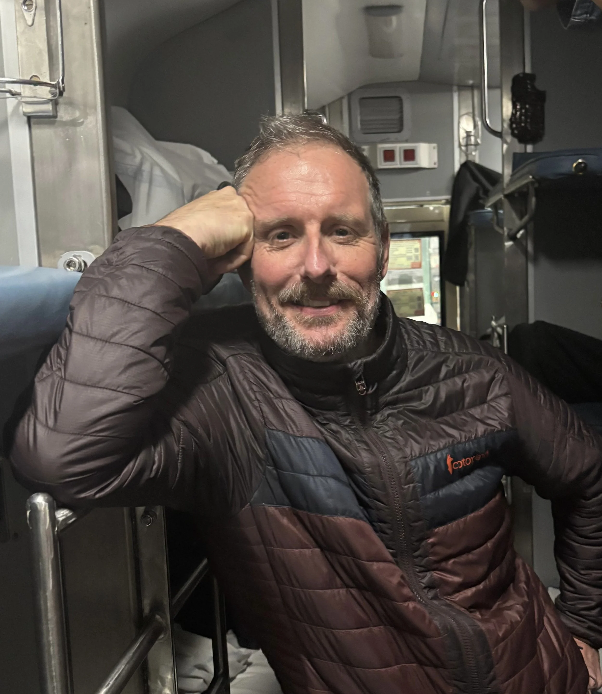 A man with short gray hair and a beard, smiling, sitting inside a vehicle or a small confined space, wearing a dark quilted jacket, resting his head on his hand.