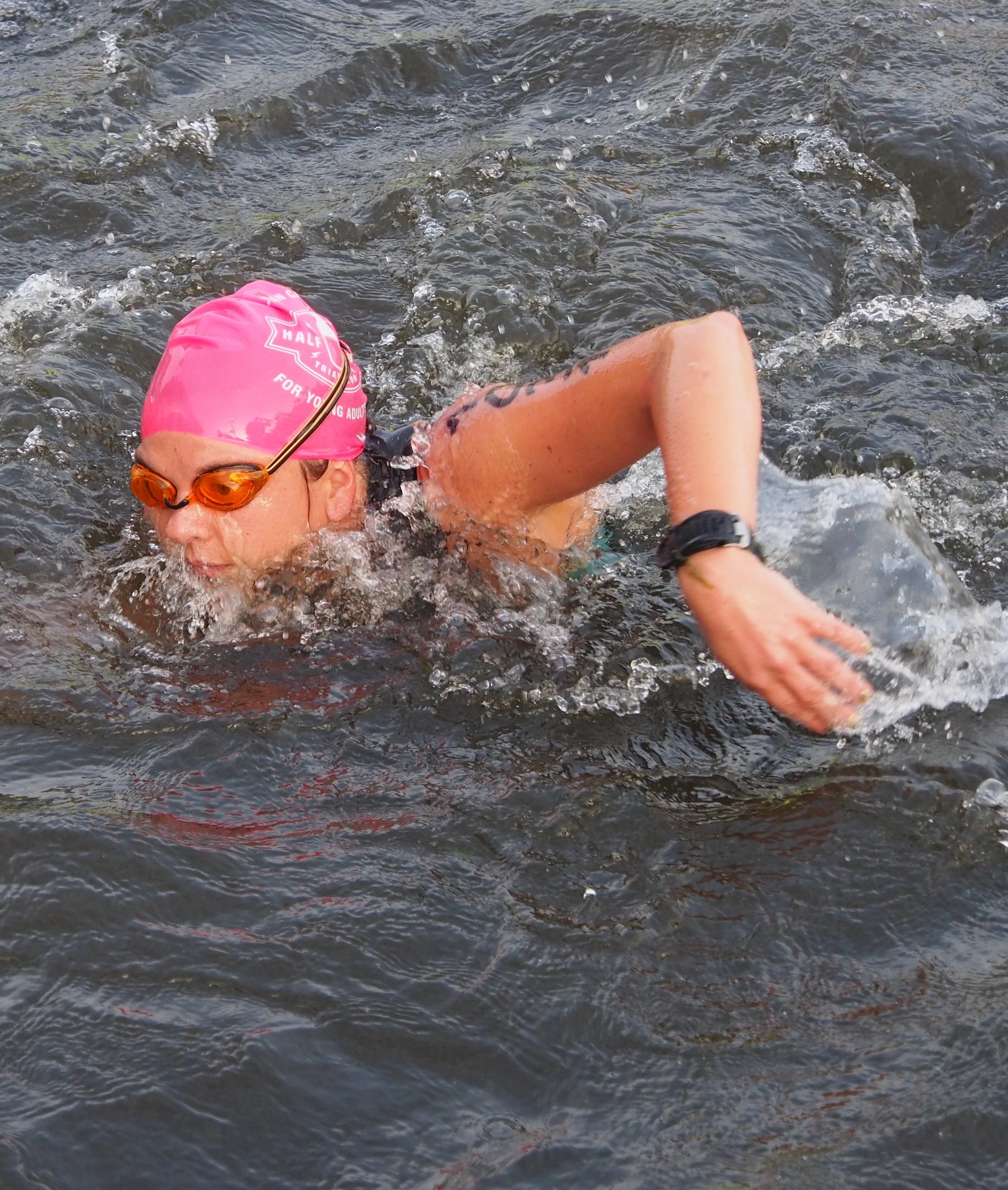 A woman wearing a pink swim cap, orange goggles, and a black watch is swimming in open water.