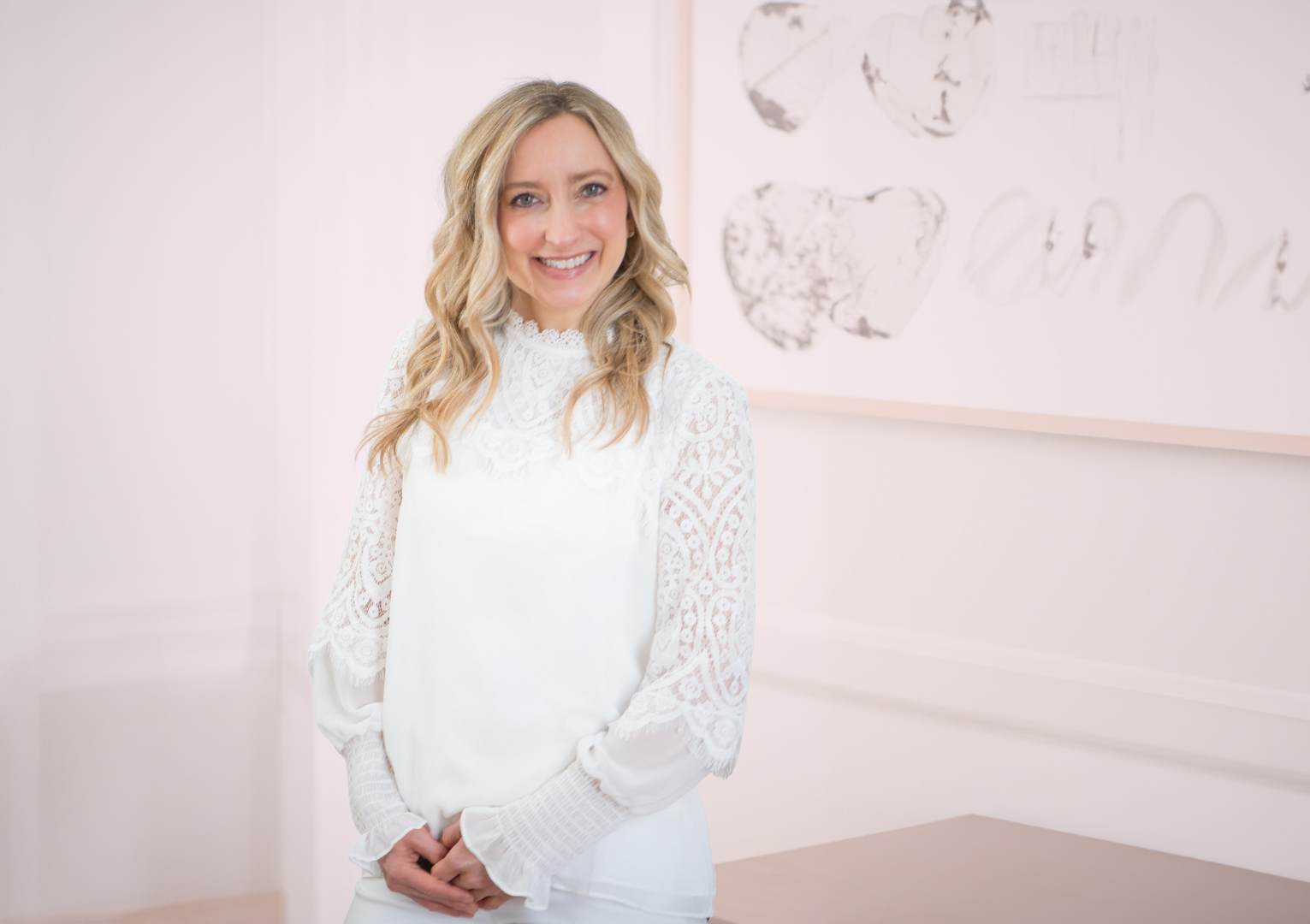 A woman with blonde wavy hair wearing a white lace blouse, standing indoors in front of abstract art on a white wall, smiling at the camera.