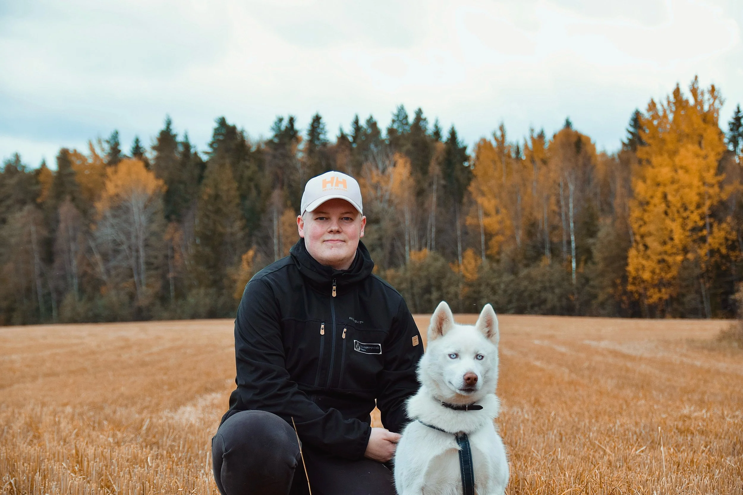 Nuori mies ja valkoinen husky-koira pellolla, taustalla syksyiset puut ja pilvinen taivas.