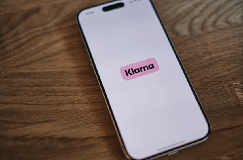 Älypuhelin, jonka näytöllä lukee 'Klarna' pinkillä taustalla, asetettuna puun pinnalle.