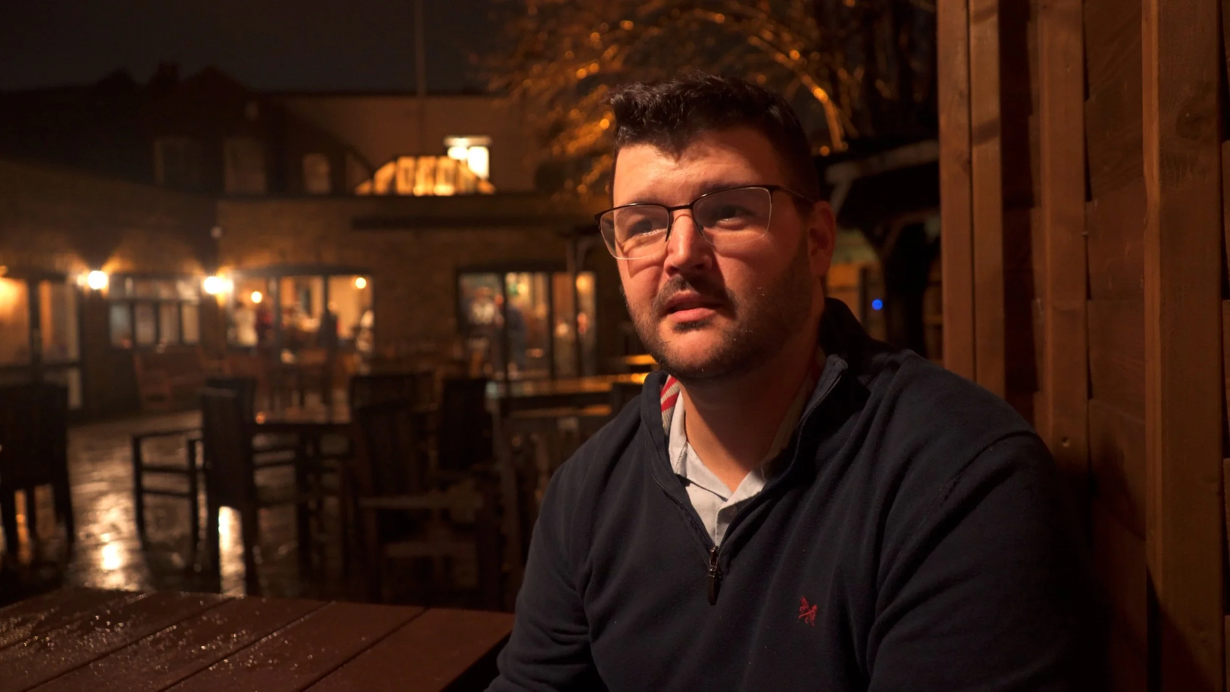 A man wearing glasses and a dark jacket sitting at a wooden table in an outdoor patio at night, illuminated by warm lighting from the background.