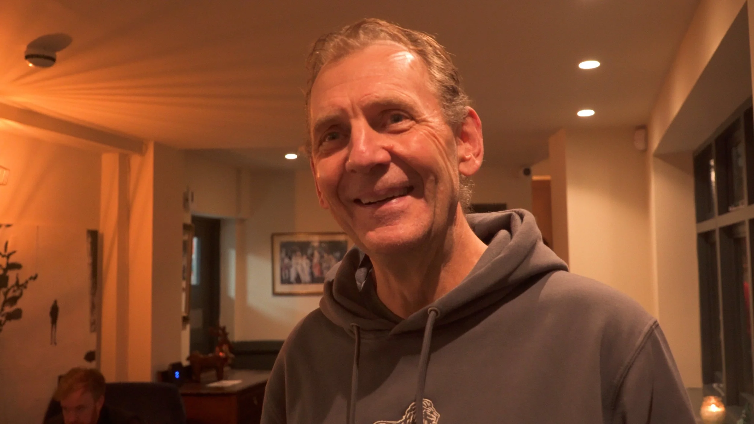 A smiling older man in a gray hoodie standing in a warmly lit room, with a framed picture and a person sitting at a desk in the background.