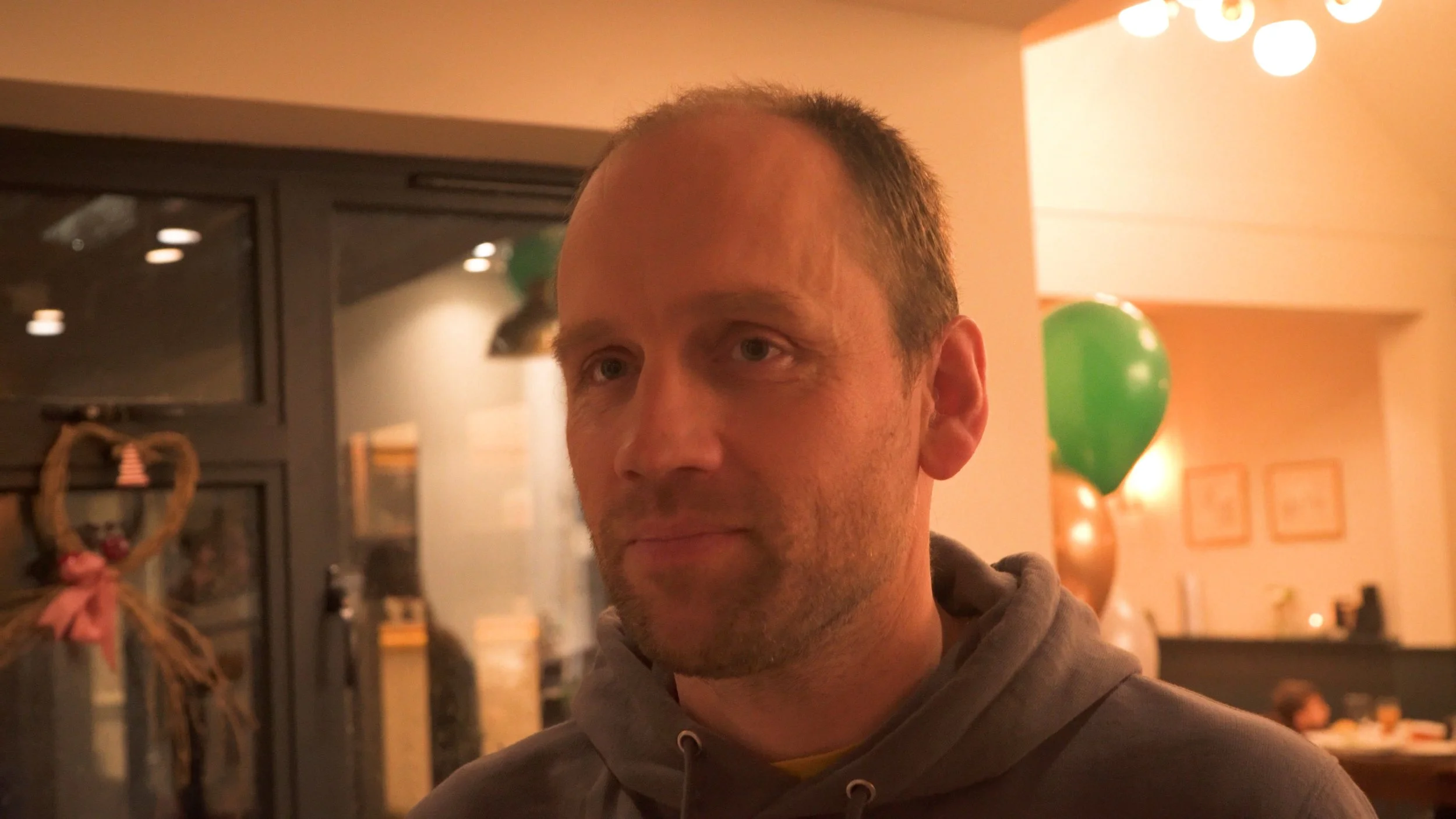 A man with short, light brown hair and wearing a brown hoodie, standing indoors at a celebration party. The background features balloons and warm lighting.