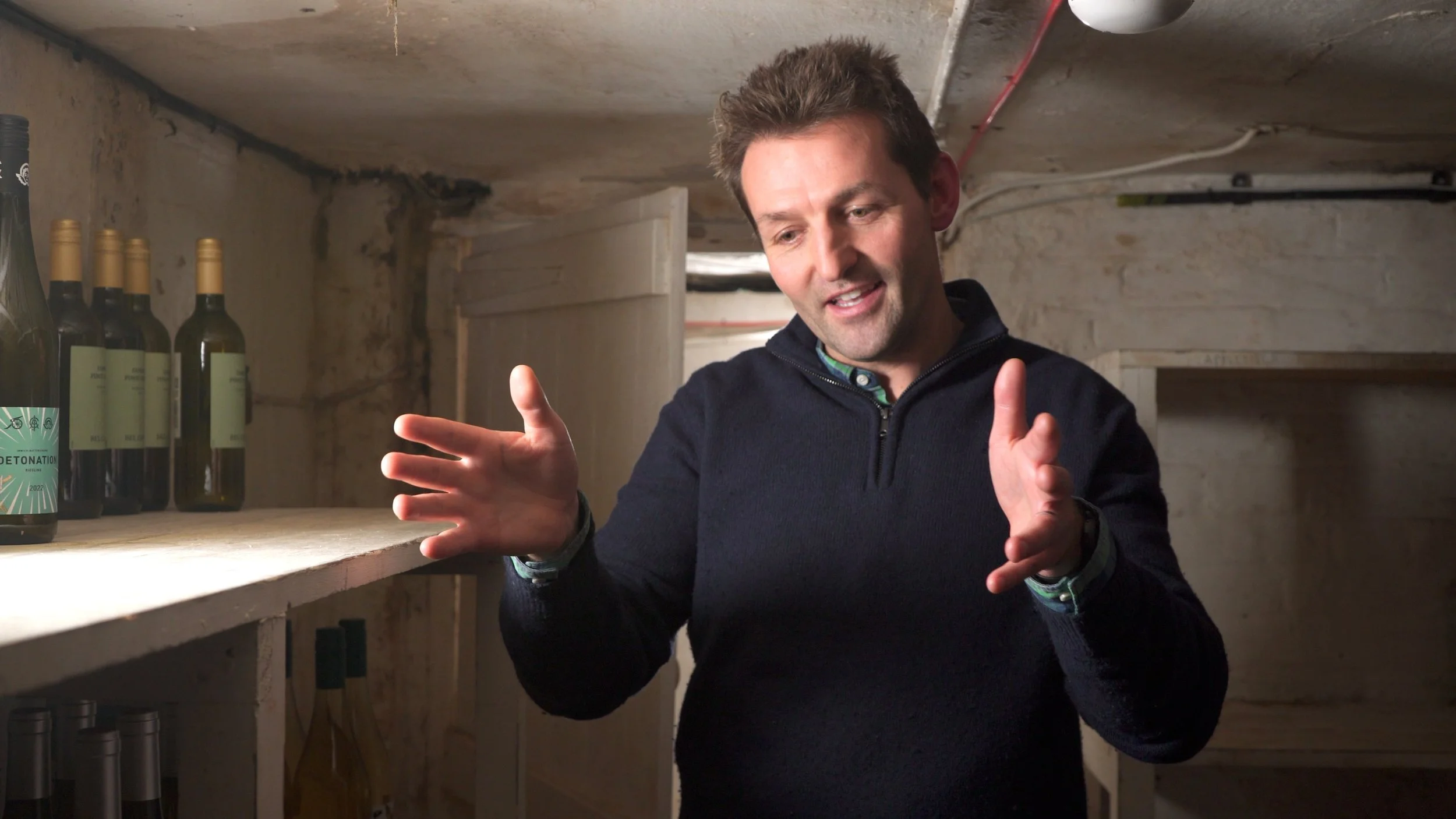 A man with brown hair wearing a dark sweater and a watch, standing in a cellar with unfinished concrete walls, gesturing with his hands, surrounded by bottles of wine on shelves.
