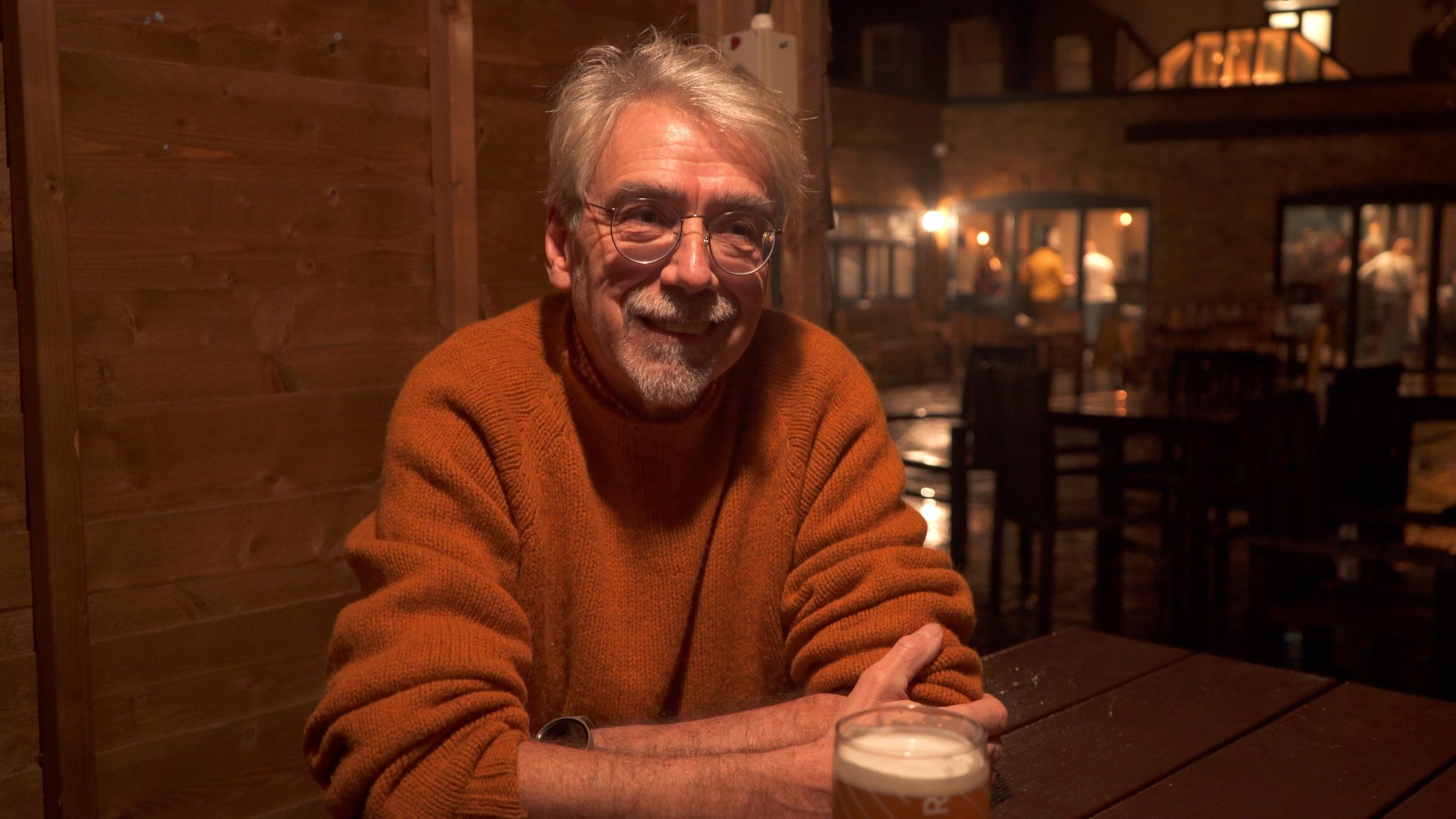 An older man with glasses and gray hair, wearing an orange sweater, smiling while sitting at a wooden table in a dimly lit bar or restaurant with wooden walls and other patrons in the background.