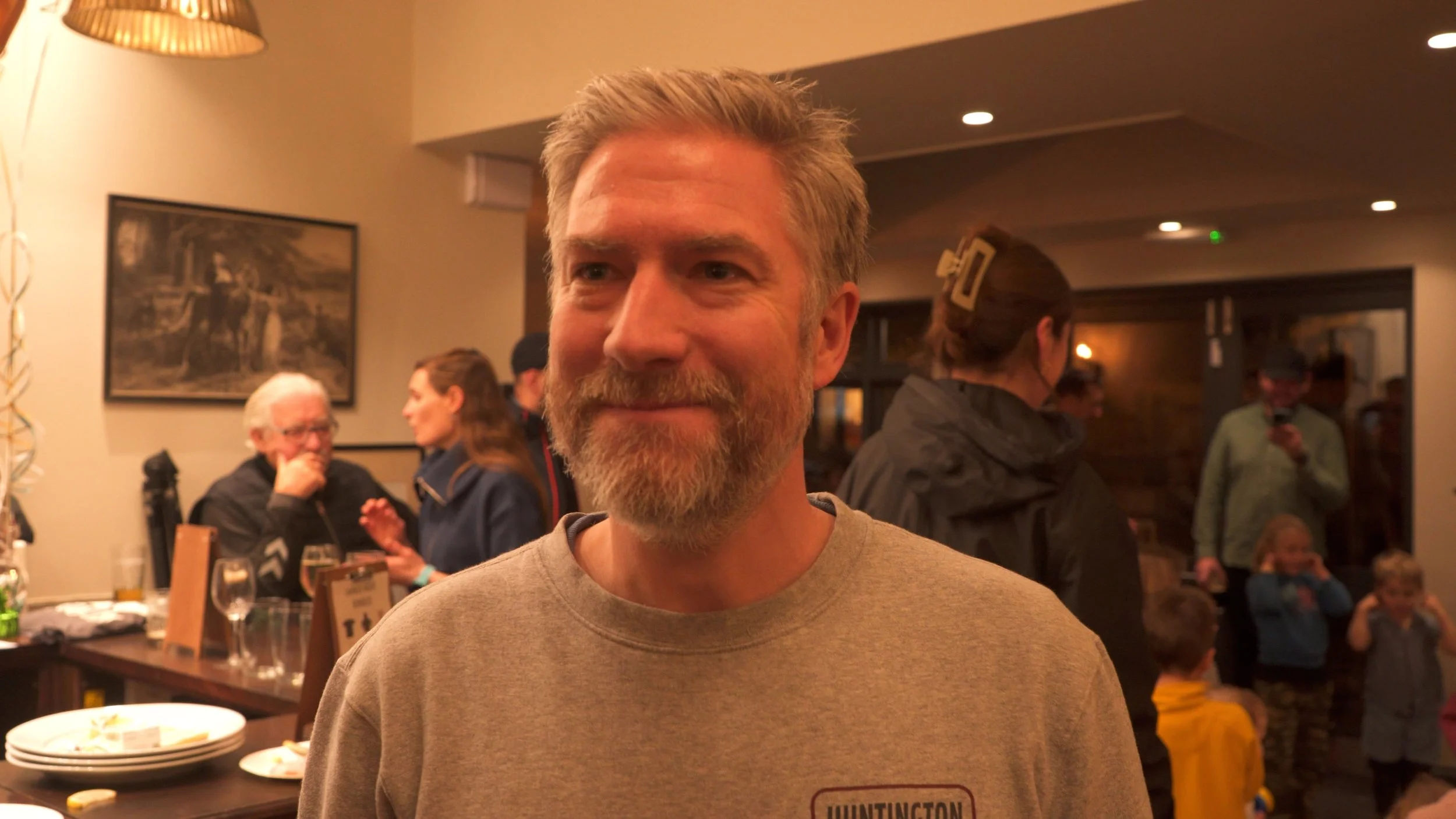 A man with a beard and mustache smiling at a social gathering in a restaurant or pub, with other people and children in the background.