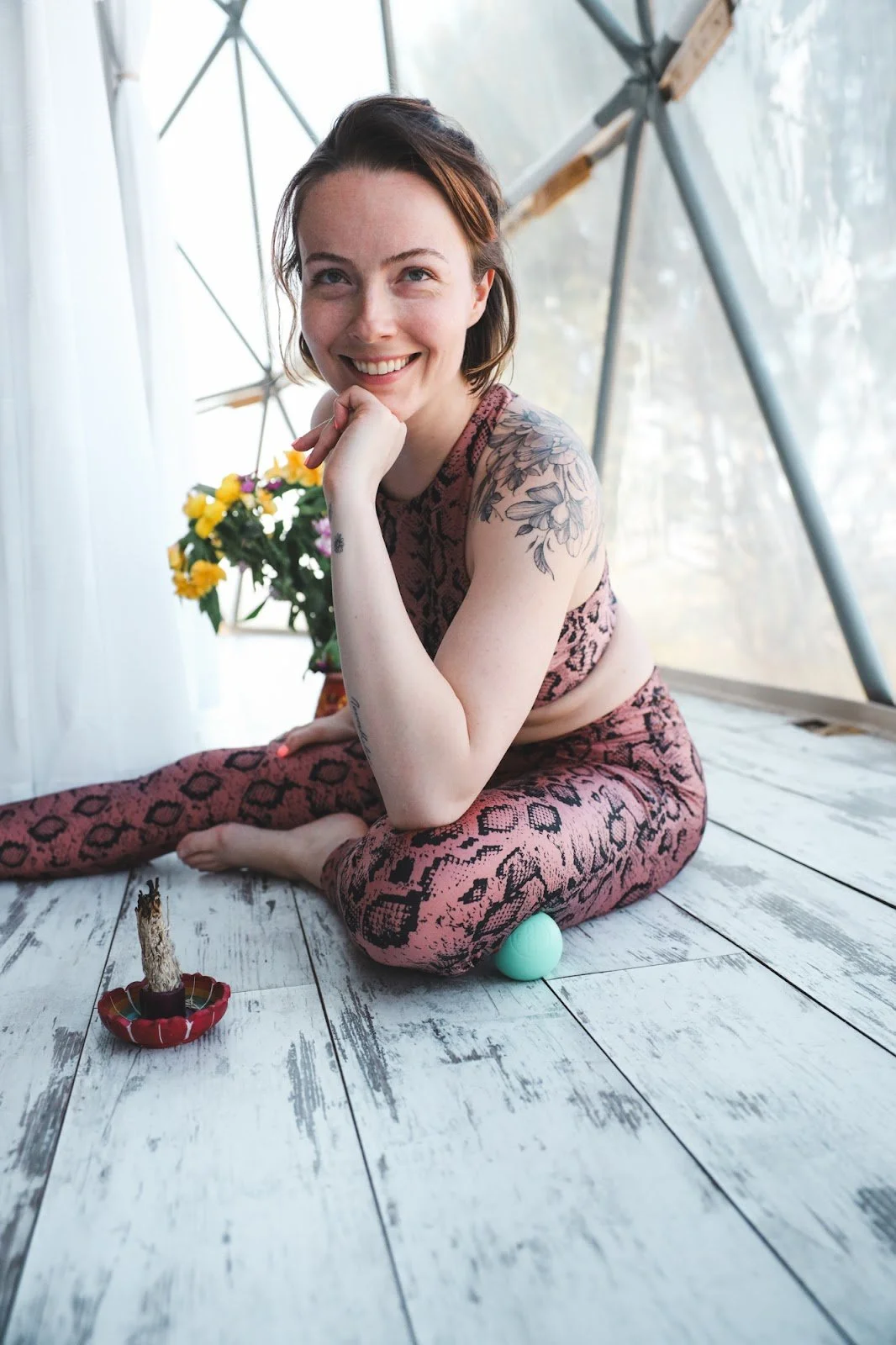 A woman with short brown hair and floral tattoos on her arm, wearing pink leopard-print yoga attire, sitting on a rustic white wooden floor inside a greenhouse or tent, smiling with her hand under her chin, with a potted plant with yellow flowers behind her and a small red dish with a wooden stick and an egg-shaped object in front of her.