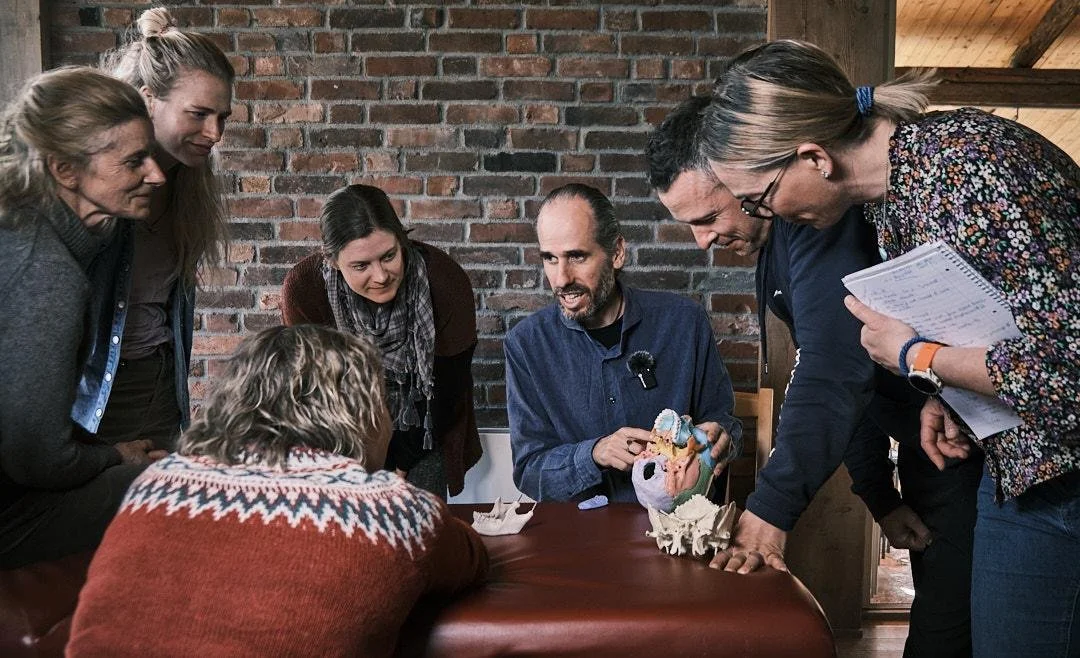 Group of six people gathered around a table, observing a model of the human heart with additional anatomical parts, in a room with a brick wall.