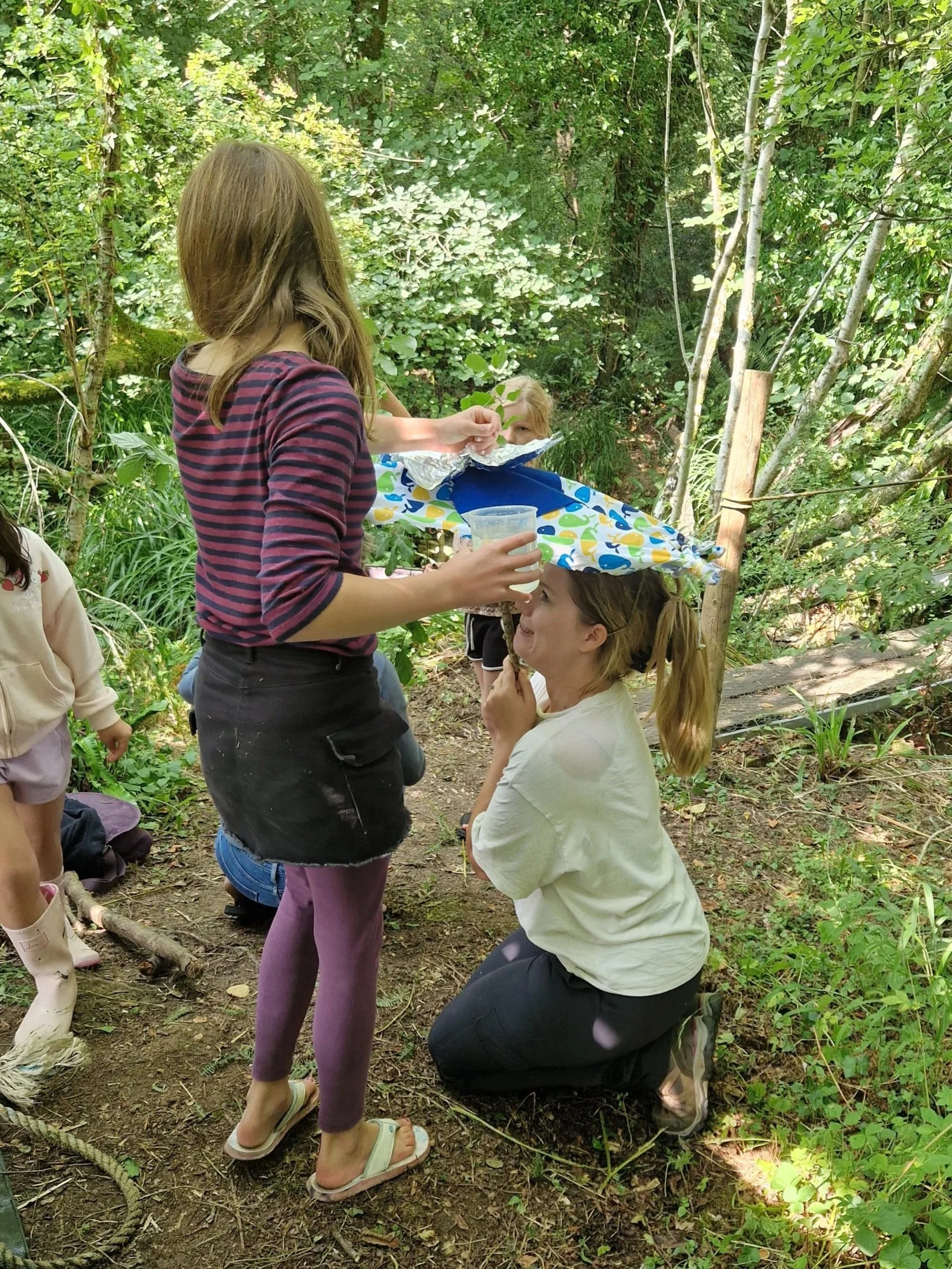 A group of children, including a girl wearing purple leggings and sandals, and a woman kneeling. Engaging in outdoor science activities.