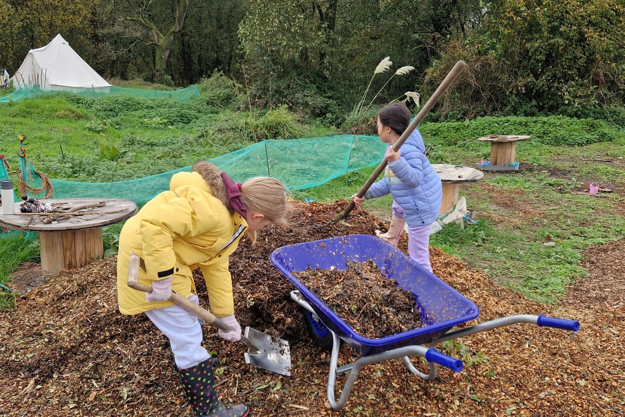Two children working together to move leaves and compost in a wheelbarrow outdoors in a garden.