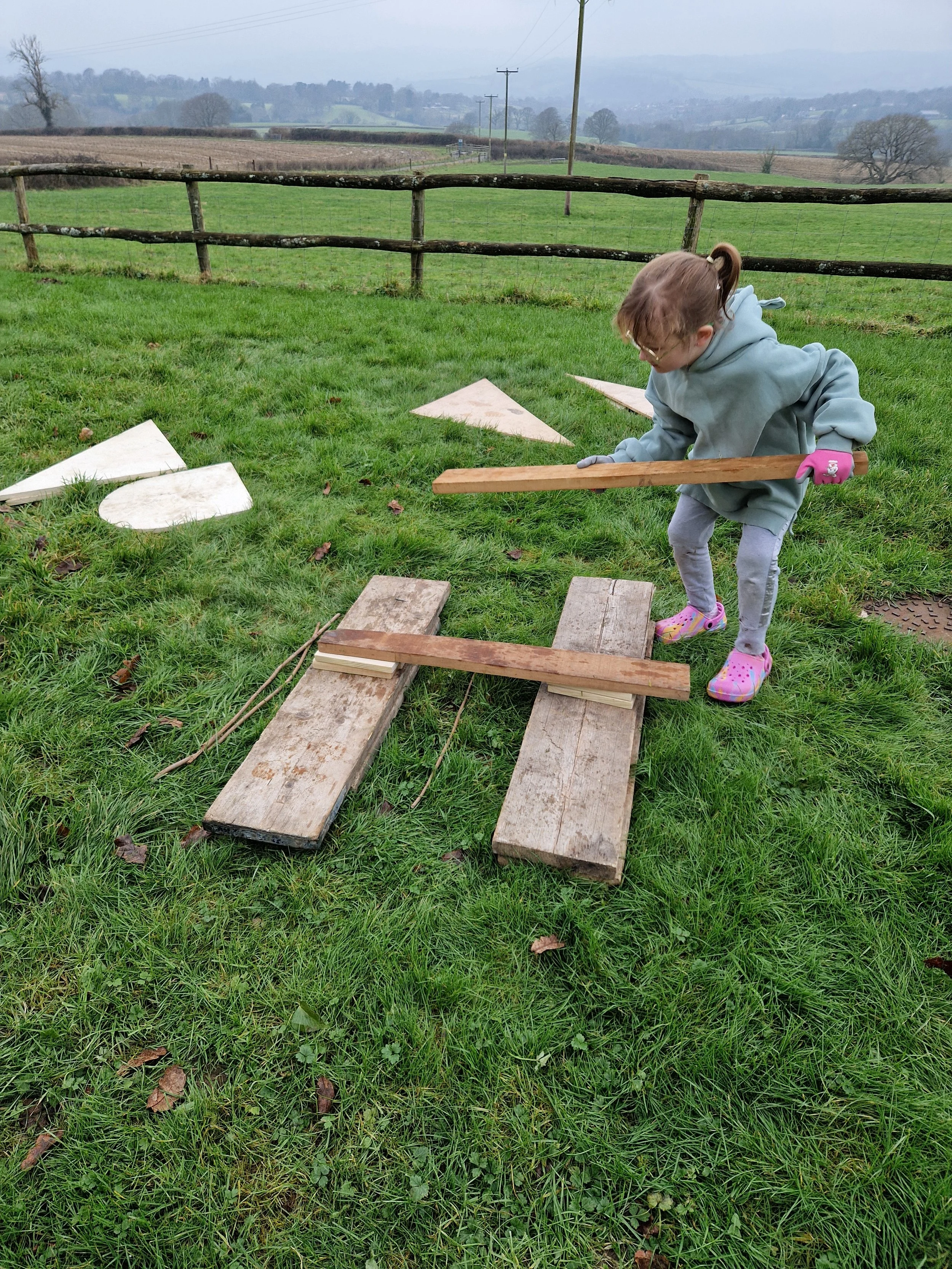A young girl is outside in a grassy field, using a wooden plank to balance on two wooden pallets placed on the ground. A rural landscape with a fence, rolling hills, and nature.