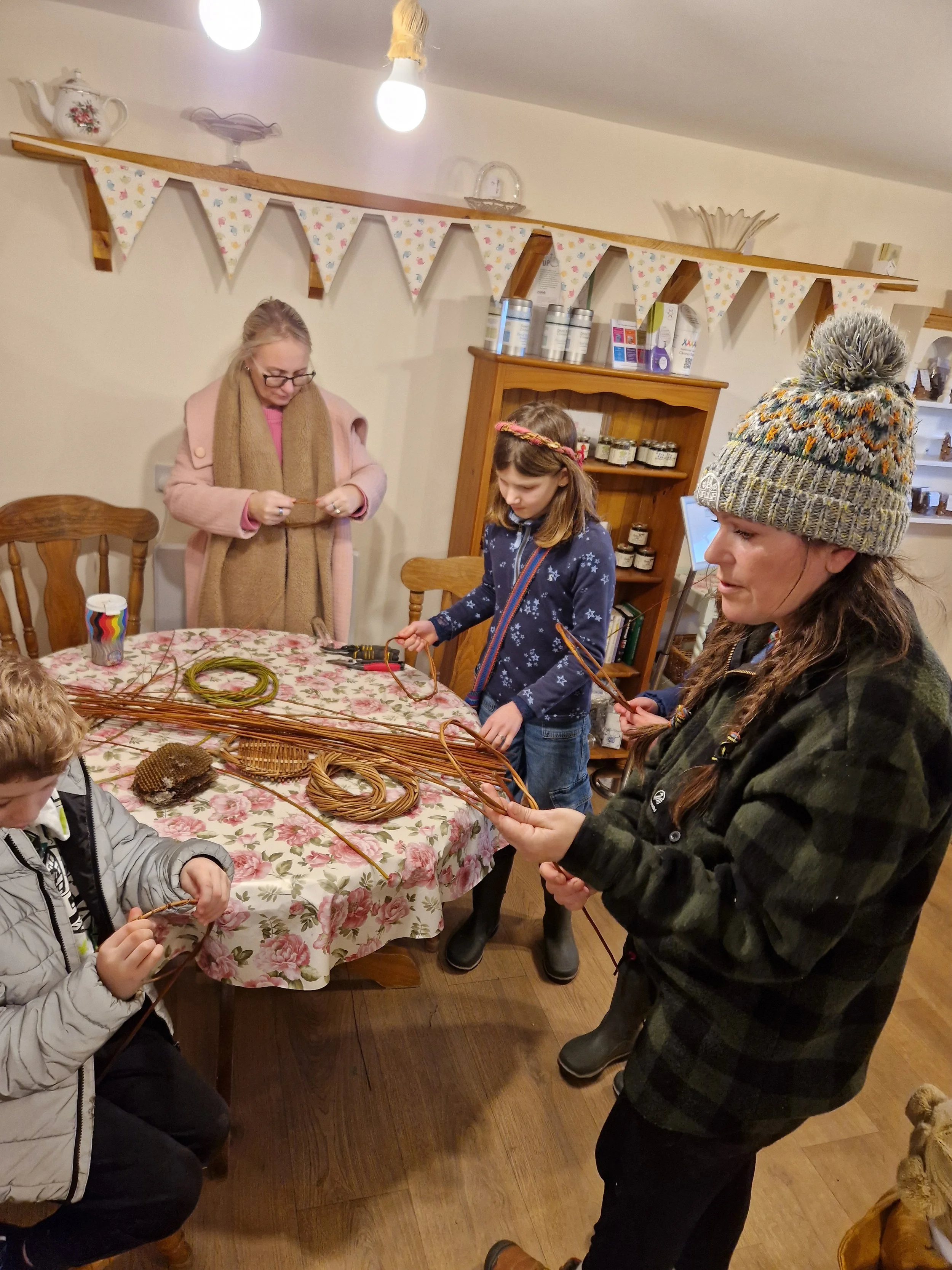A group of children and an adult working with basketry materials around a dining table in a cozy home setting.