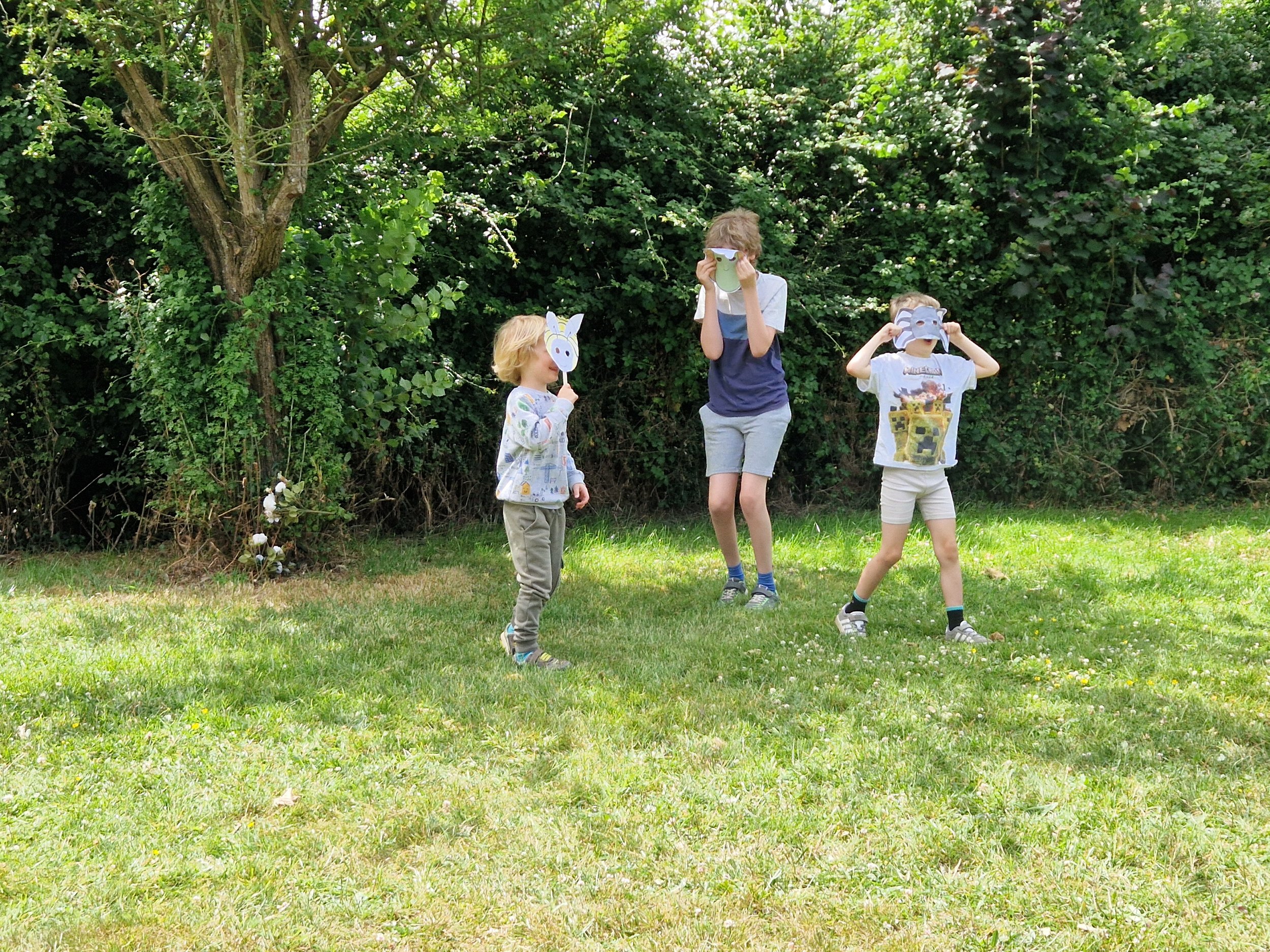 Three children wearing animal masks playing in a grassy yard with trees and bushes in the background.