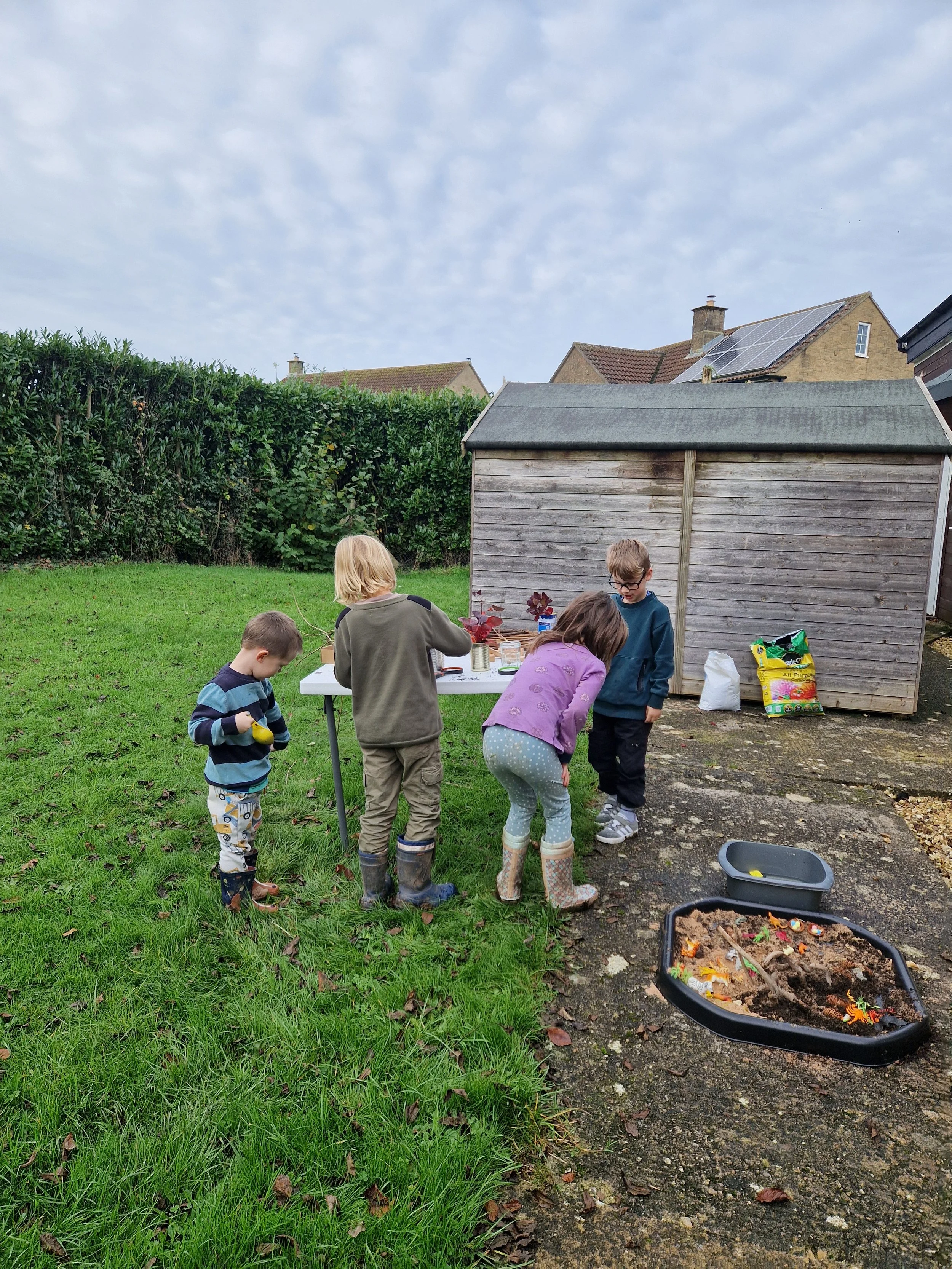 Four children playing outside near a shed and a table, with tools and gardening supplies, on a cloudy day.
