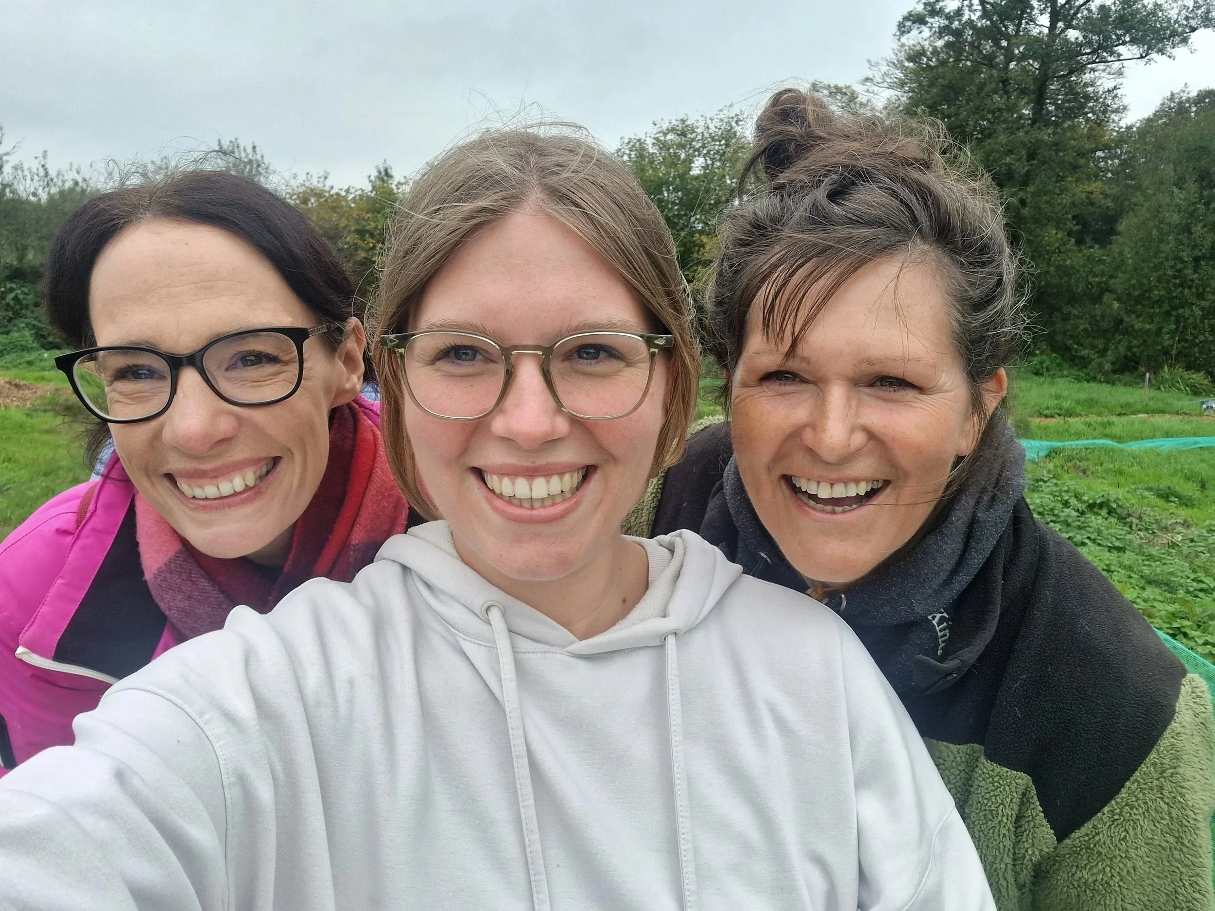 Three women smiling outdoors in a green garden with trees in the background.