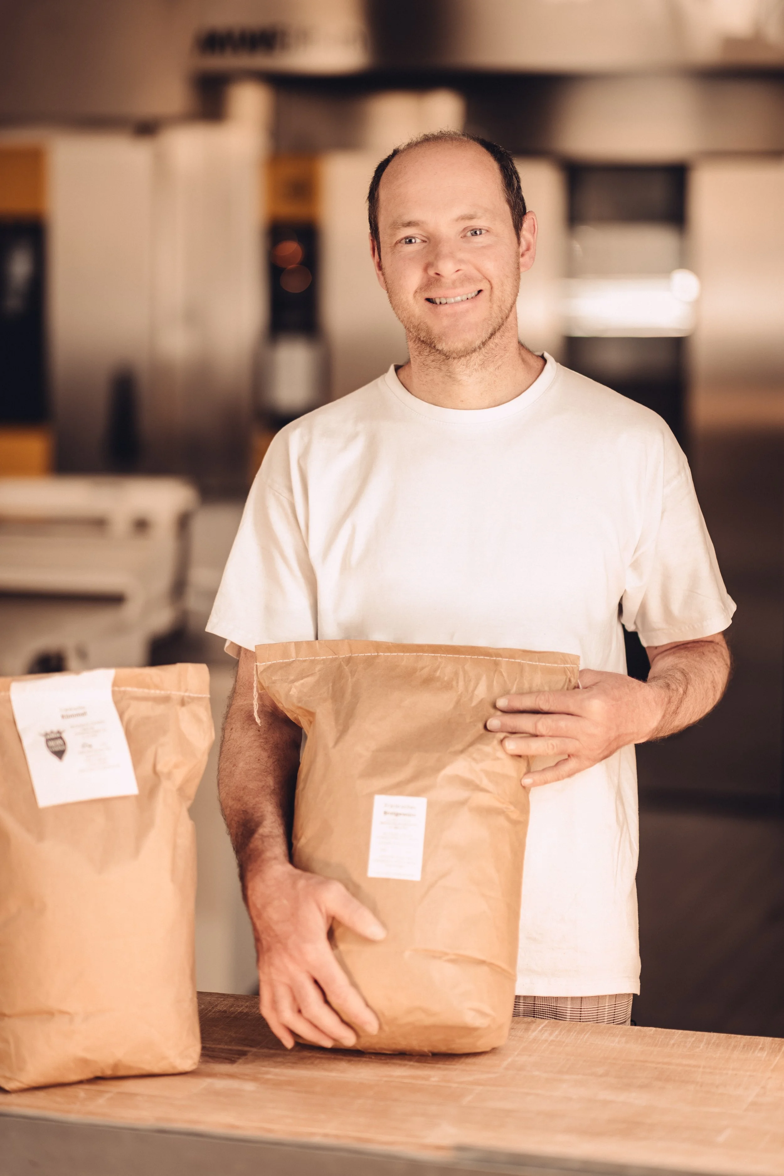 Ein lachender Mann in weißem T-Shirt hält eine braune Papiertüte in den Händen, im Hintergrund sind weitere Papiertüten auf einem Tisch. Es sieht nach einer Verpackungsarbeit aus.
