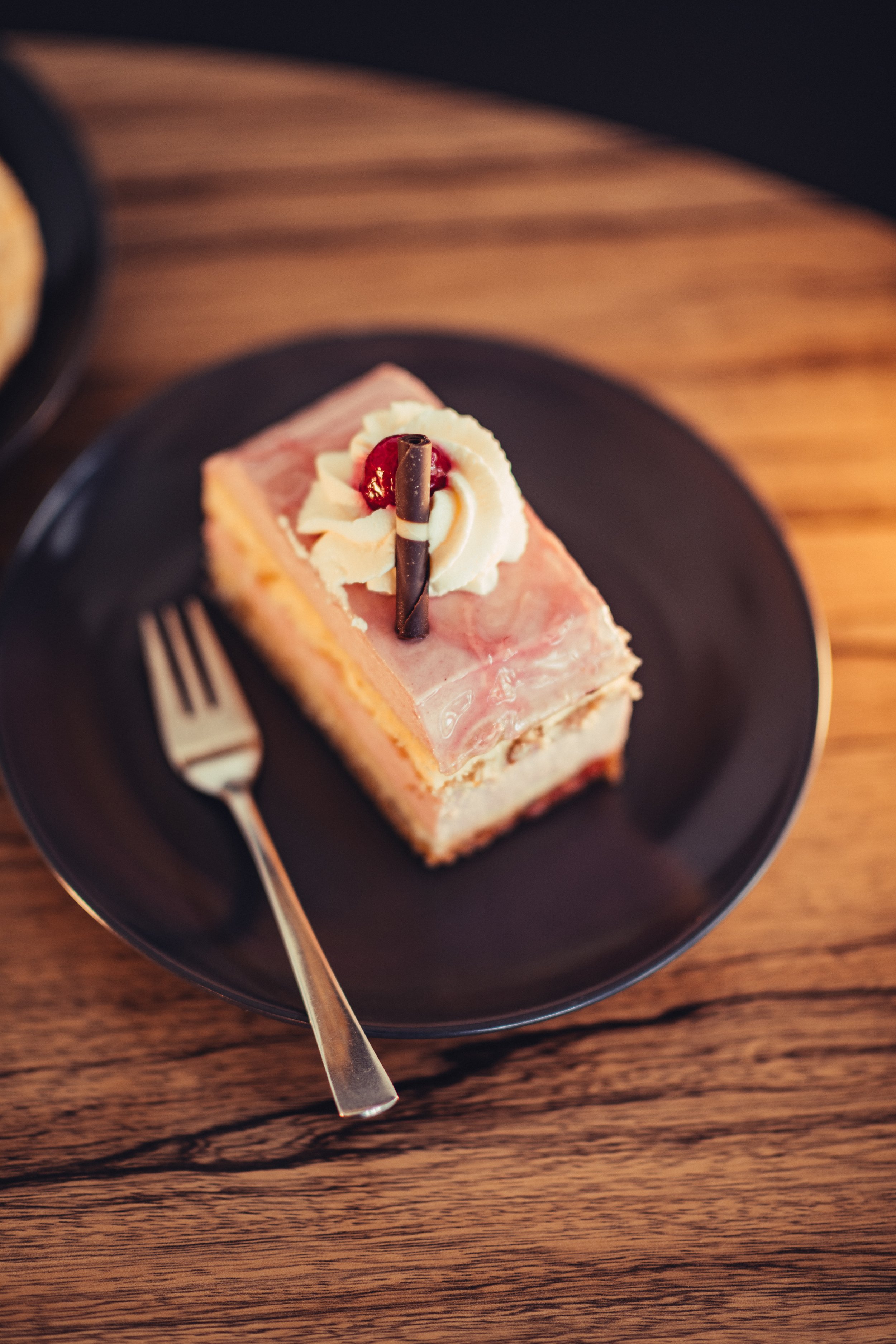 Ein Stück Kuchen mit weißer Glasur, Kirsch und Schokoladenstange auf einem schwarzen Teller, auf einem Holztisch, mit Gabel daneben.