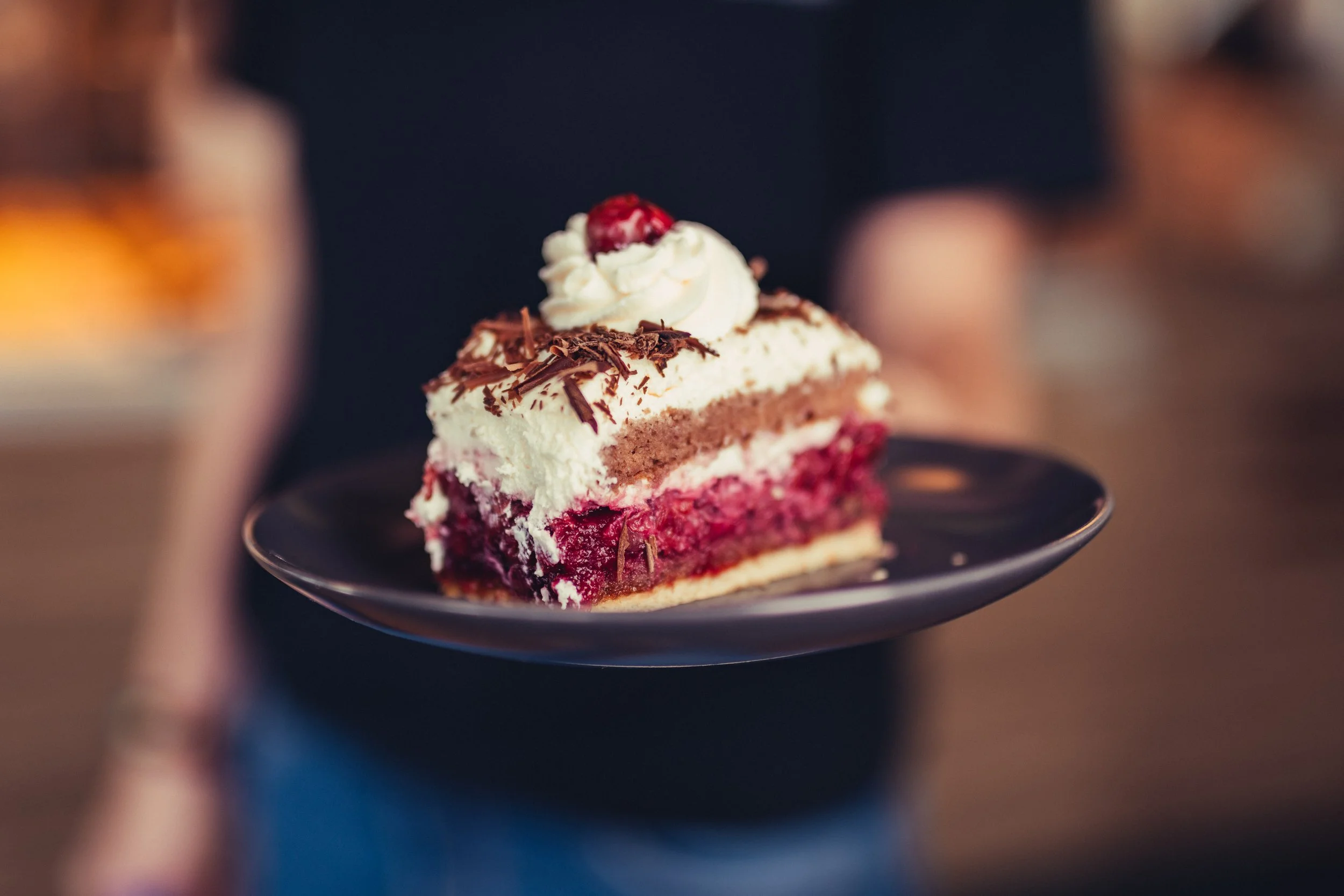 Ein Stück Erdbeerkuchen mit Sahne, Schokoladenraspeln und einer Kirsche auf einem schwarzen Teller, mit einem unscharfen Hintergrund.