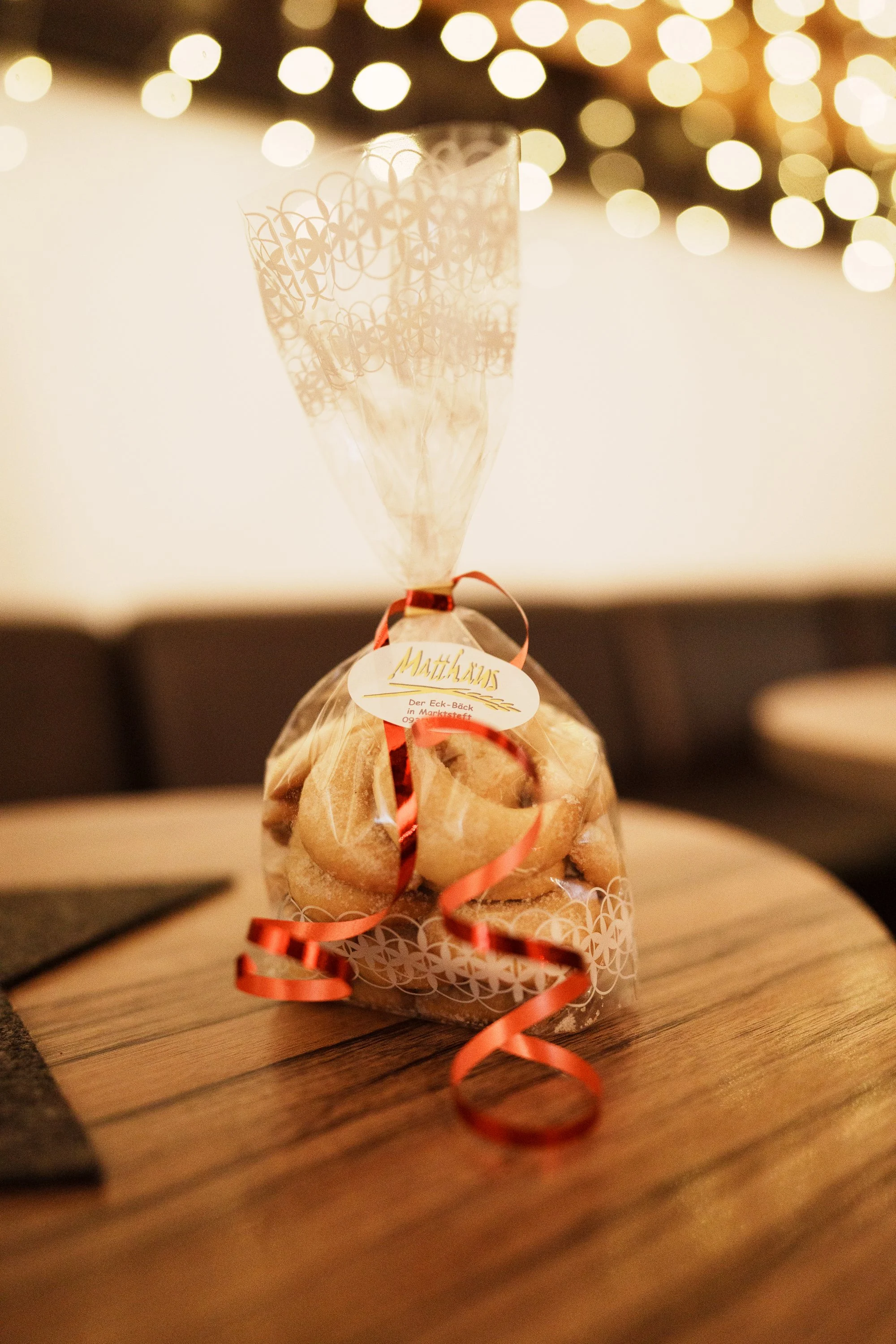 Ein Mitbringsel aus Bäckerei, verpackt in transparentem Geschenkpapier mit roten Schleifen, steht auf einem Holztisch, im Hintergrund Lichter im Bokeh-Effekt.