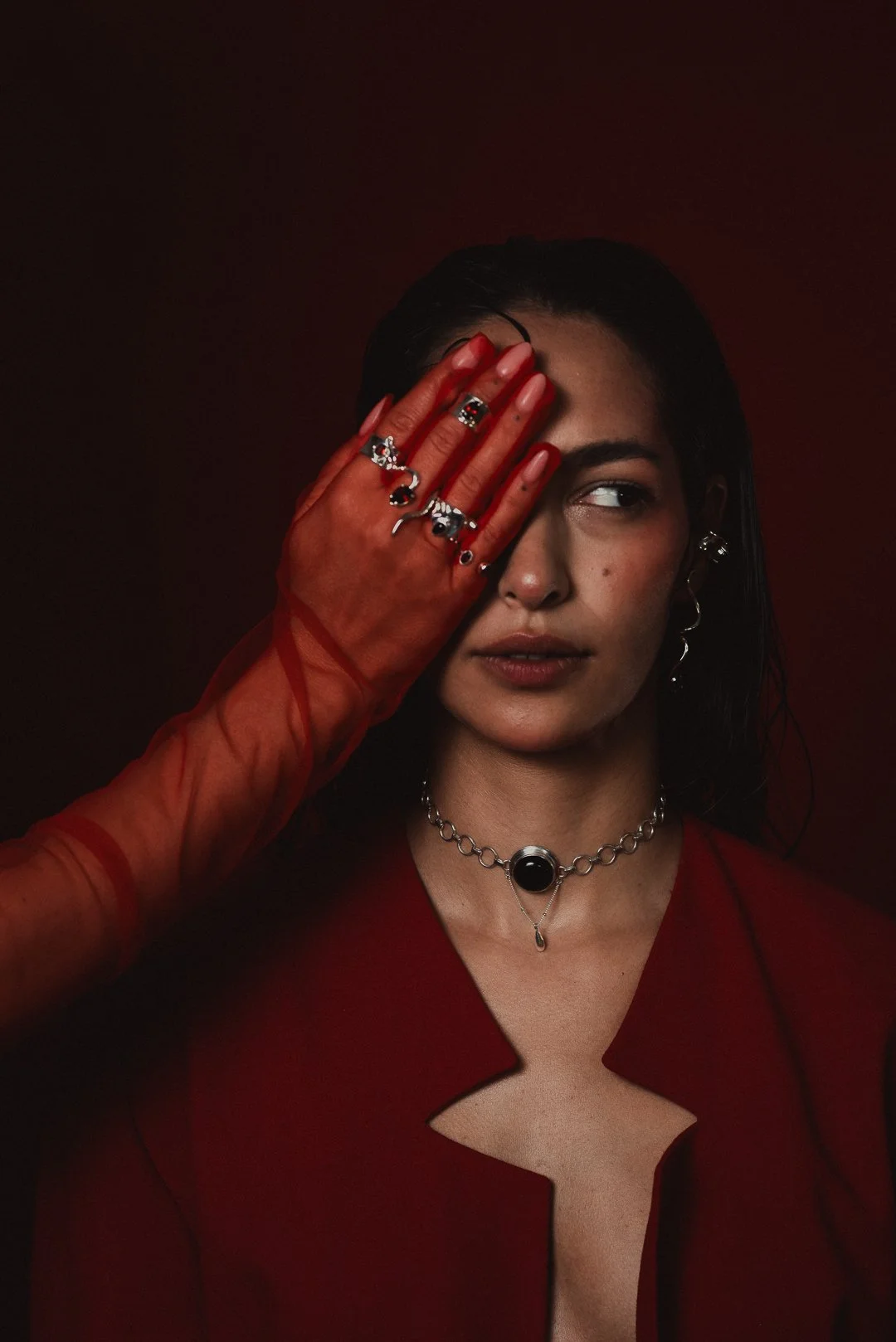 Mujer con piel clara, teñida de rojo y maquillaje natural, sosteniendo su cara con la mano izquierda, usando guantes rojos y varias joyas plateadas, con fondo oscuro y vestimenta roja.