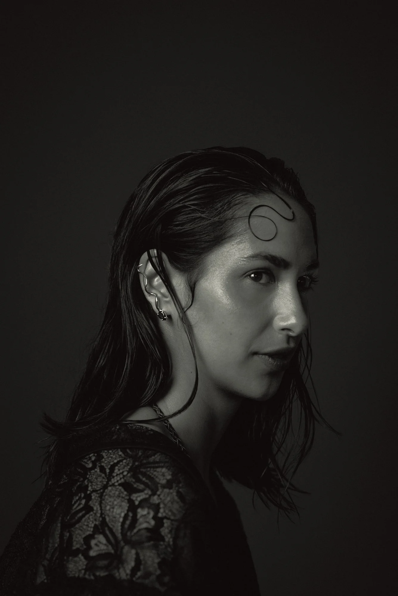 Retrato en blanco y negro de una mujer con cabello oscuro y rizado, con el rostro en perfil, vestida con un top de encaje y usando varios pendientes.